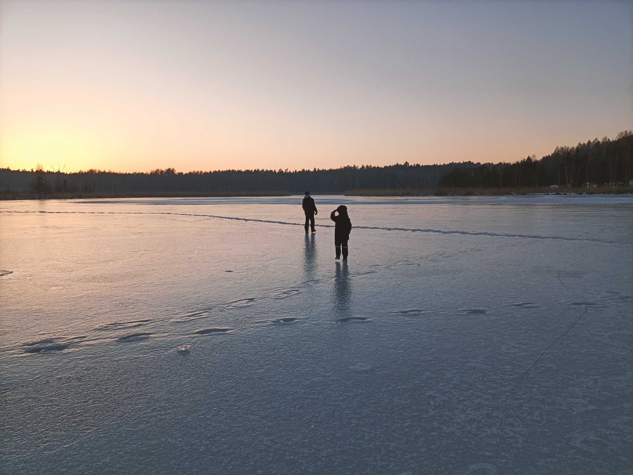 Dwie osoby spacerują po zamarzniętym jeziorze o zmierzchu. Ślady na lodzie prowadzą w stronę lasu na horyzoncie, niebo w odcieniach różu i pomarańczy.