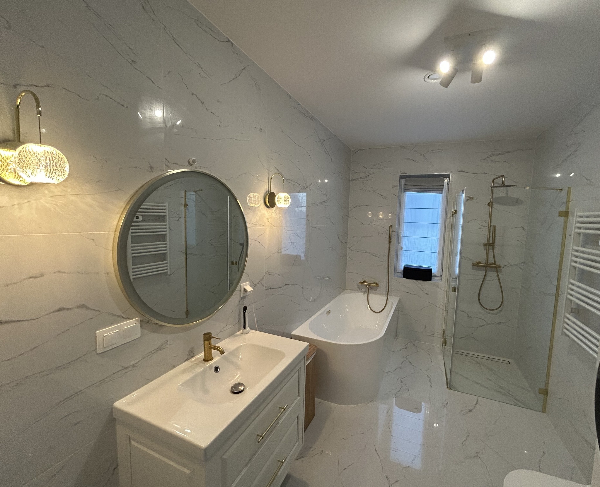 Elegancka łazienka w bieli i złocie: marmurowe płytki, okrągłe lustro, wolnostojąca wanna i kabina prysznicowa ze złotymi akcentami. Nowoczesne oświetlenie.