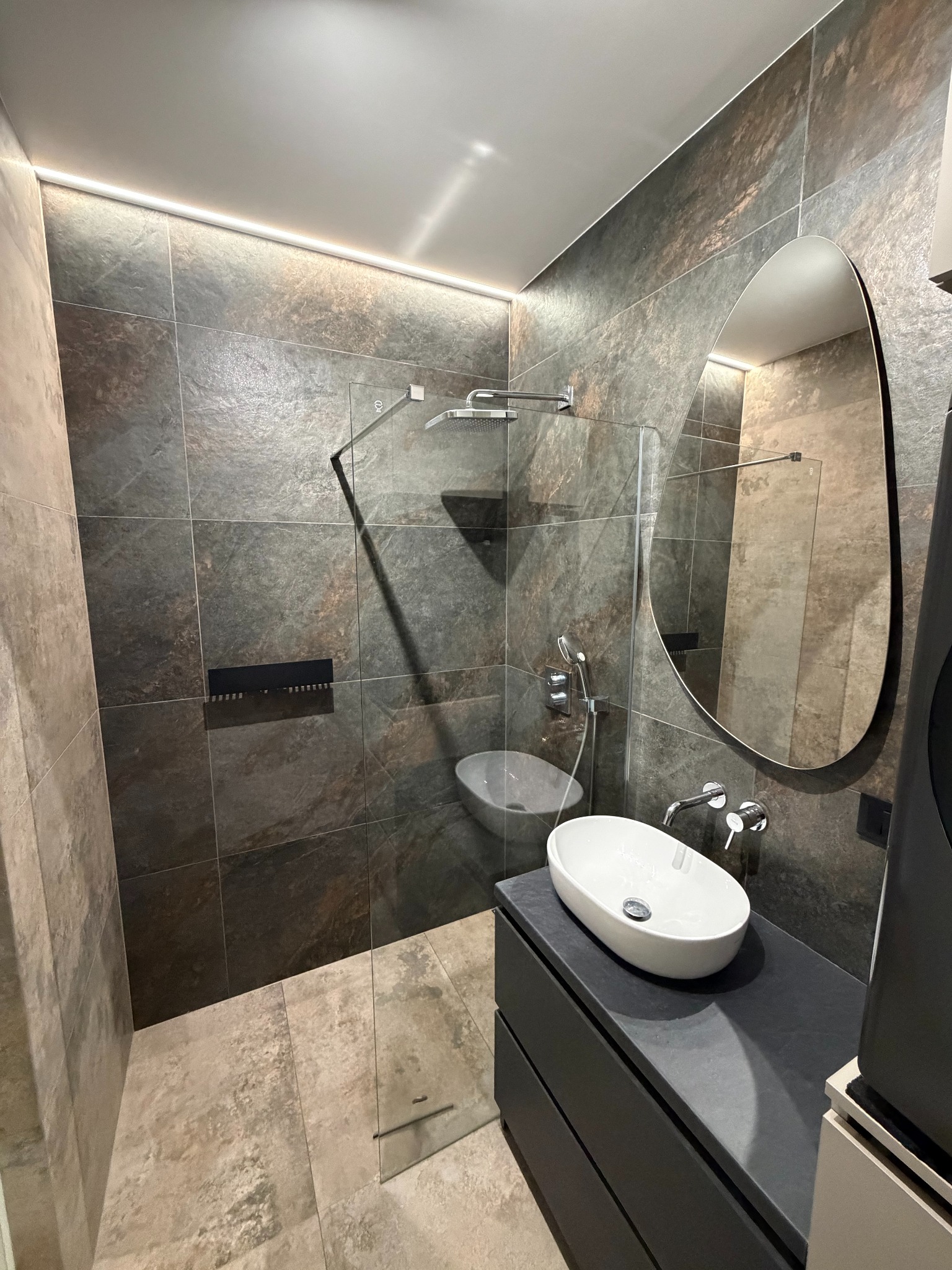 Nowoczesna łazienka z kabiną prysznicową typu walk-in, umywalką nablatową na ciemnej szafce i owalnym lustrem. Ciemne płytki imitujące kamień na ścianach i podłodze.