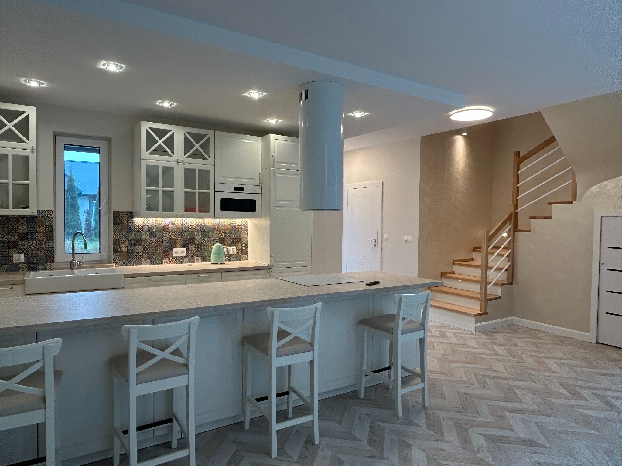 Jasna kuchnia z wyspą i hokerami, widok na schody. Białe meble, wzorzysta mozaika nad blatem. Parkiet ułożony w jodełkę. Nowoczesny design.