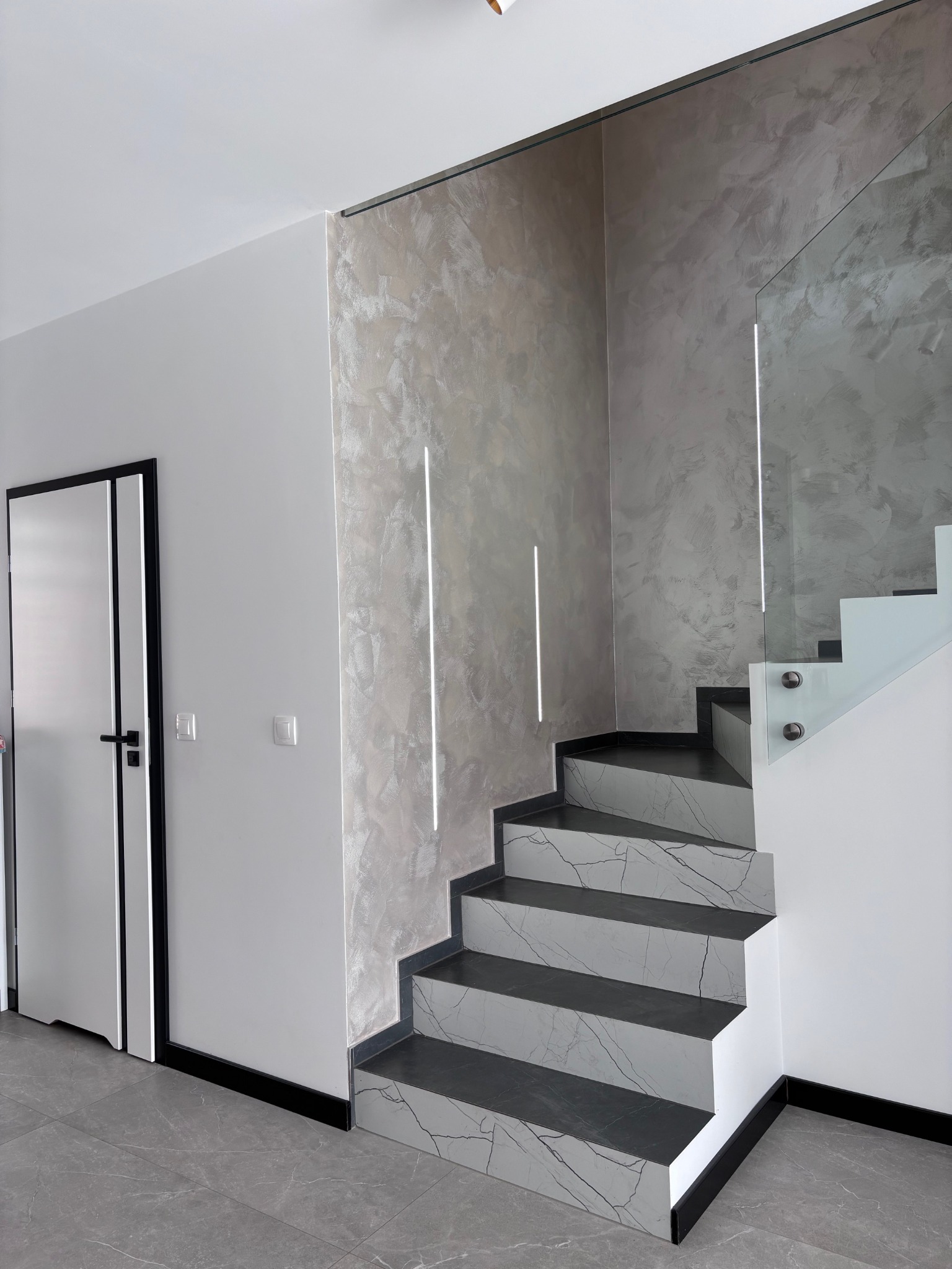 Nowoczesne schody z marmurowym wzorem i szklaną balustradą, oświetlone pionowymi lampami na ścianie z dekoracyjnym tynkiem. Widoczne drzwi z czarną ramą i szara podłoga.