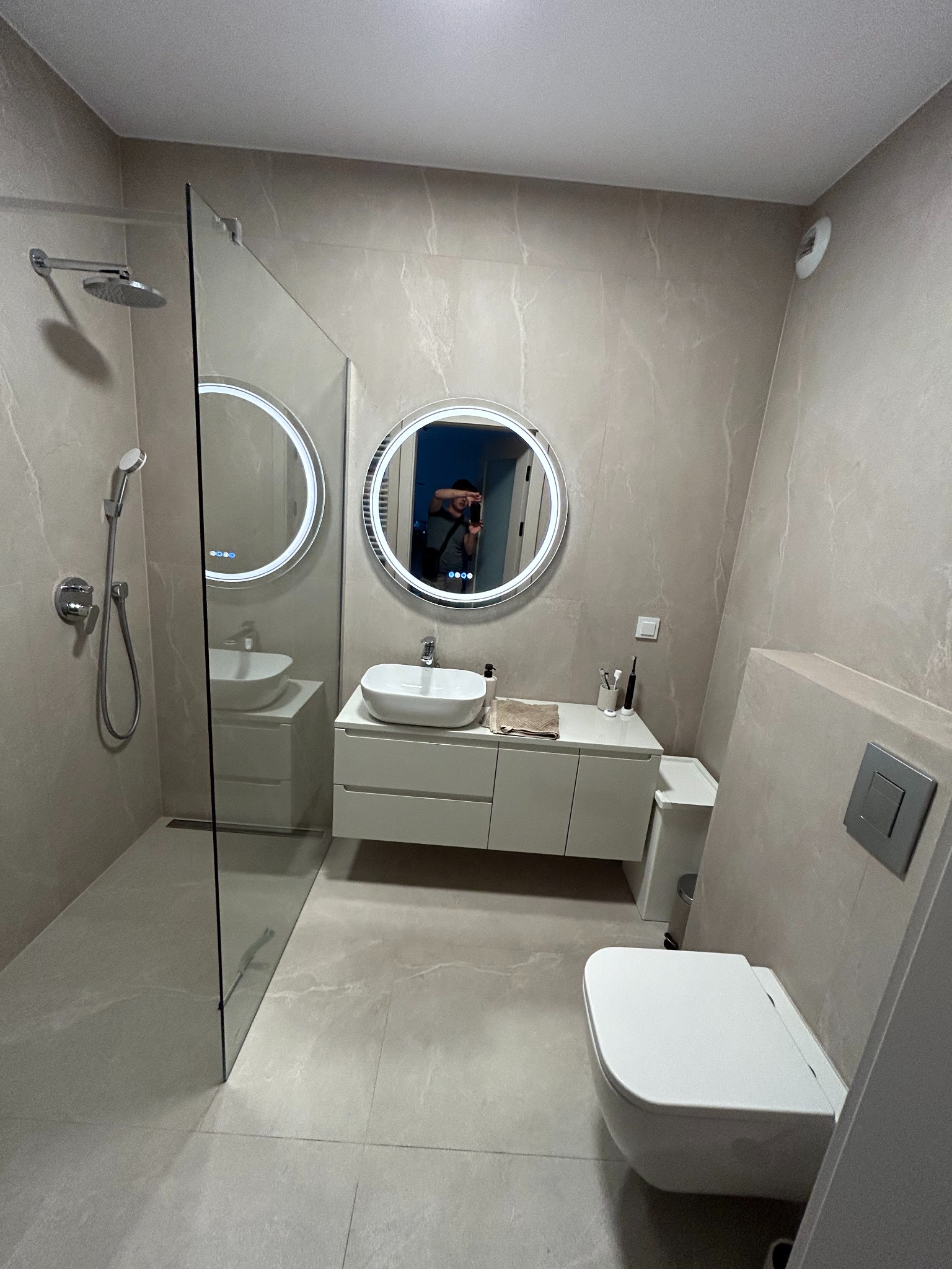 Nowoczesna łazienka z kabiną prysznicową typu walk-in, podświetlanym lustrem i wiszącą toaletą. Stonowana kolorystyka, płytki imitujące marmur, minimalistyczny design.