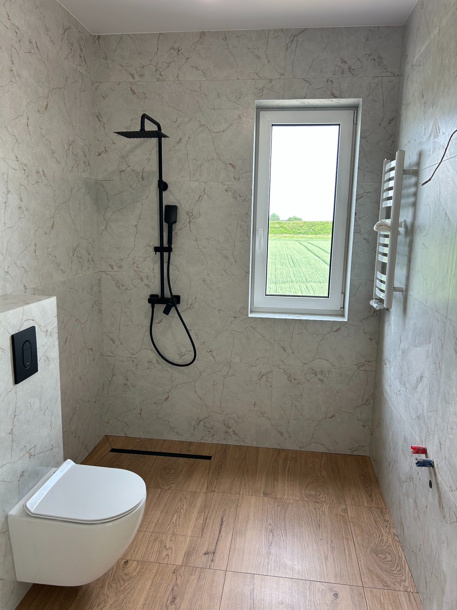 Nowoczesna łazienka z marmurowymi płytkami, czarną armaturą prysznicową i drewnopodobną podłogą. Widok na zielone pole przez okno.