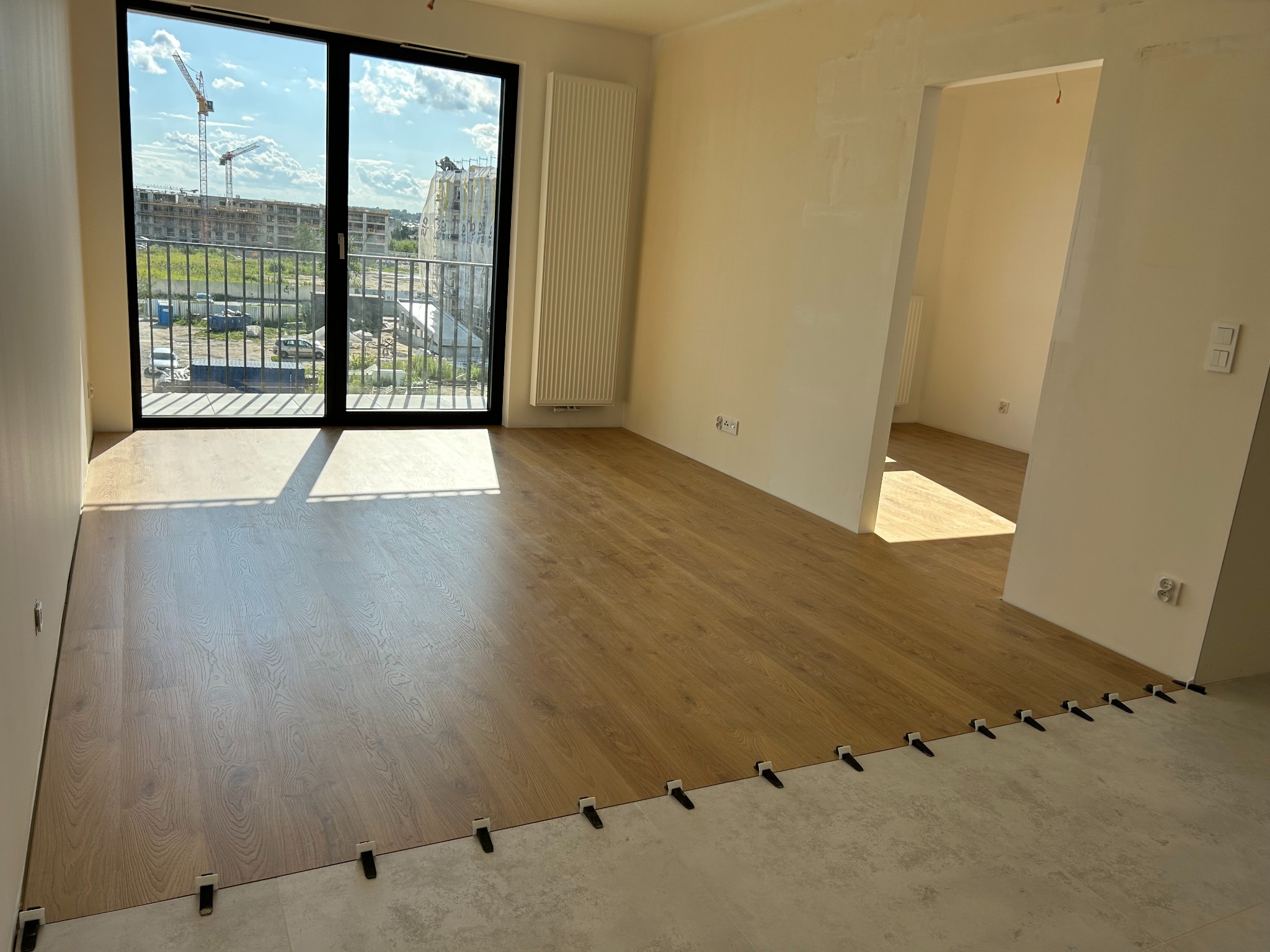 Układanie paneli podłogowych w jasnym wnętrzu z widokiem na budowę za oknem. Panele w odcieniach brązu, widoczne czarne kliny dystansowe.