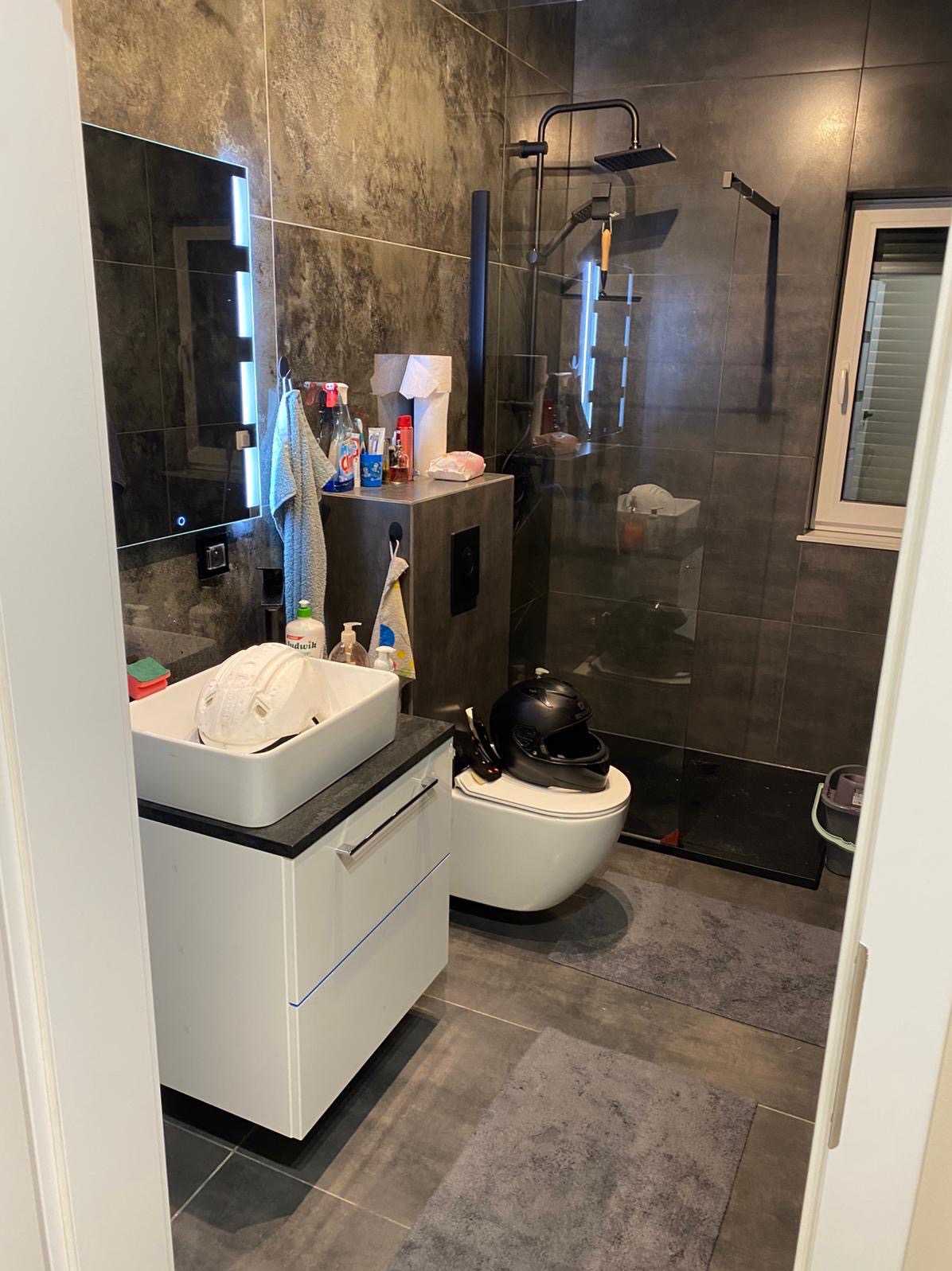 Nowoczesna łazienka z szarymi płytkami, kabiną prysznicową typu walk-in i białą szafką umywalkową. Widoczny kask na toalecie i akcesoria łazienkowe.