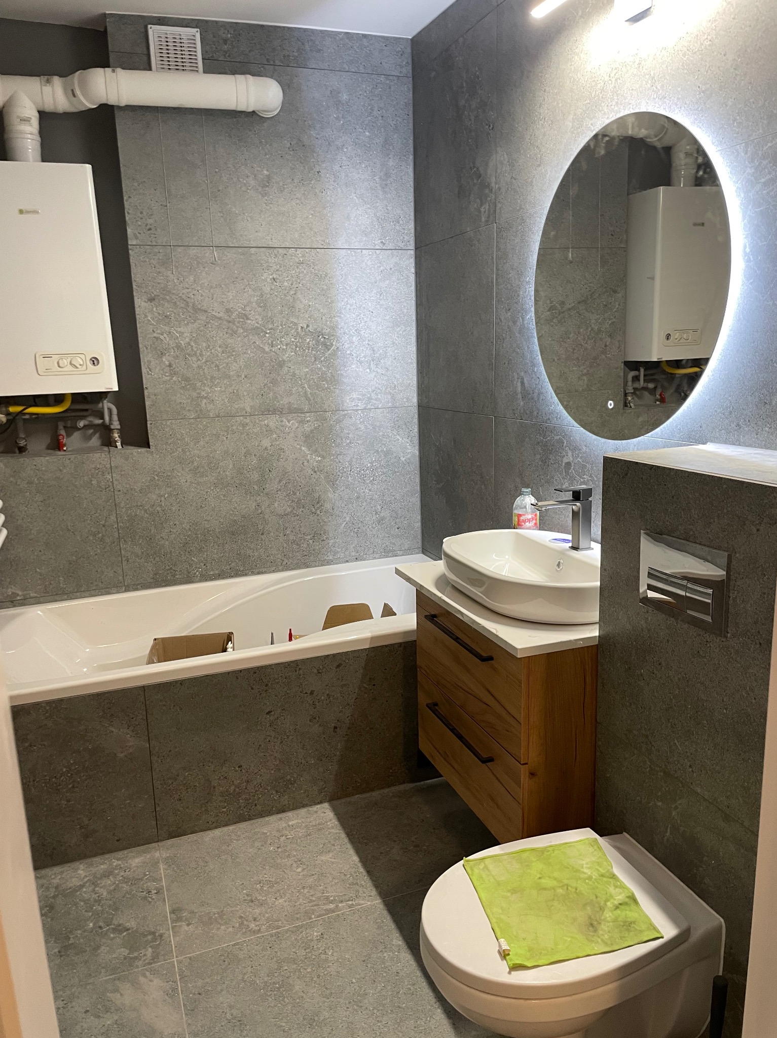 Wykończona łazienka w szarościach z okrągłym lustrem podświetlanym LED, drewnianą szafką pod umywalką i wiszącą toaletą. Na sedesie leży zielona ściereczka.