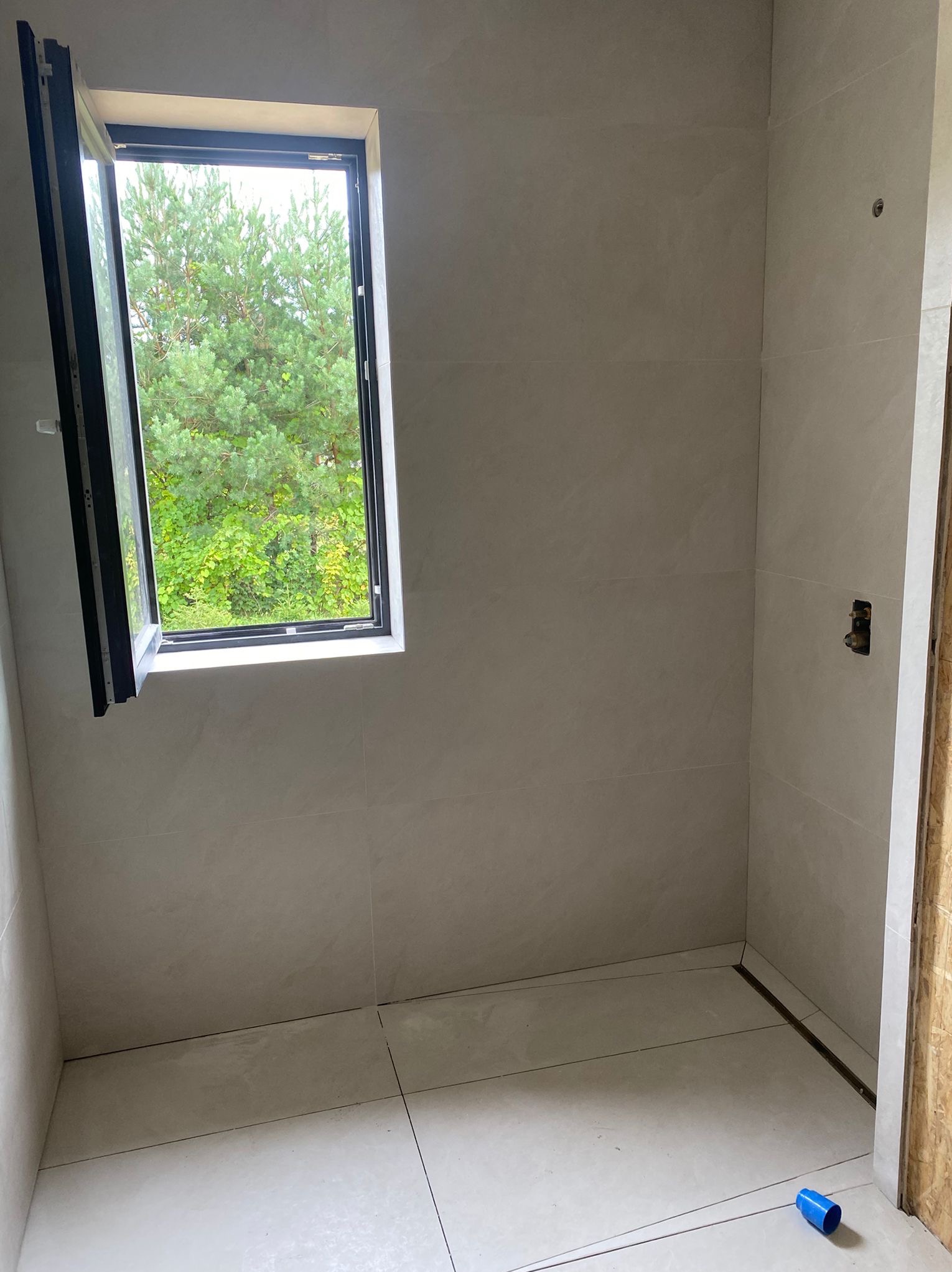 Wykończona łazienka z jasnymi płytkami, otwartym oknem z widokiem na zieleń. Widoczne detale instalacji i niebieski element na podłodze. Minimalistyczny design.