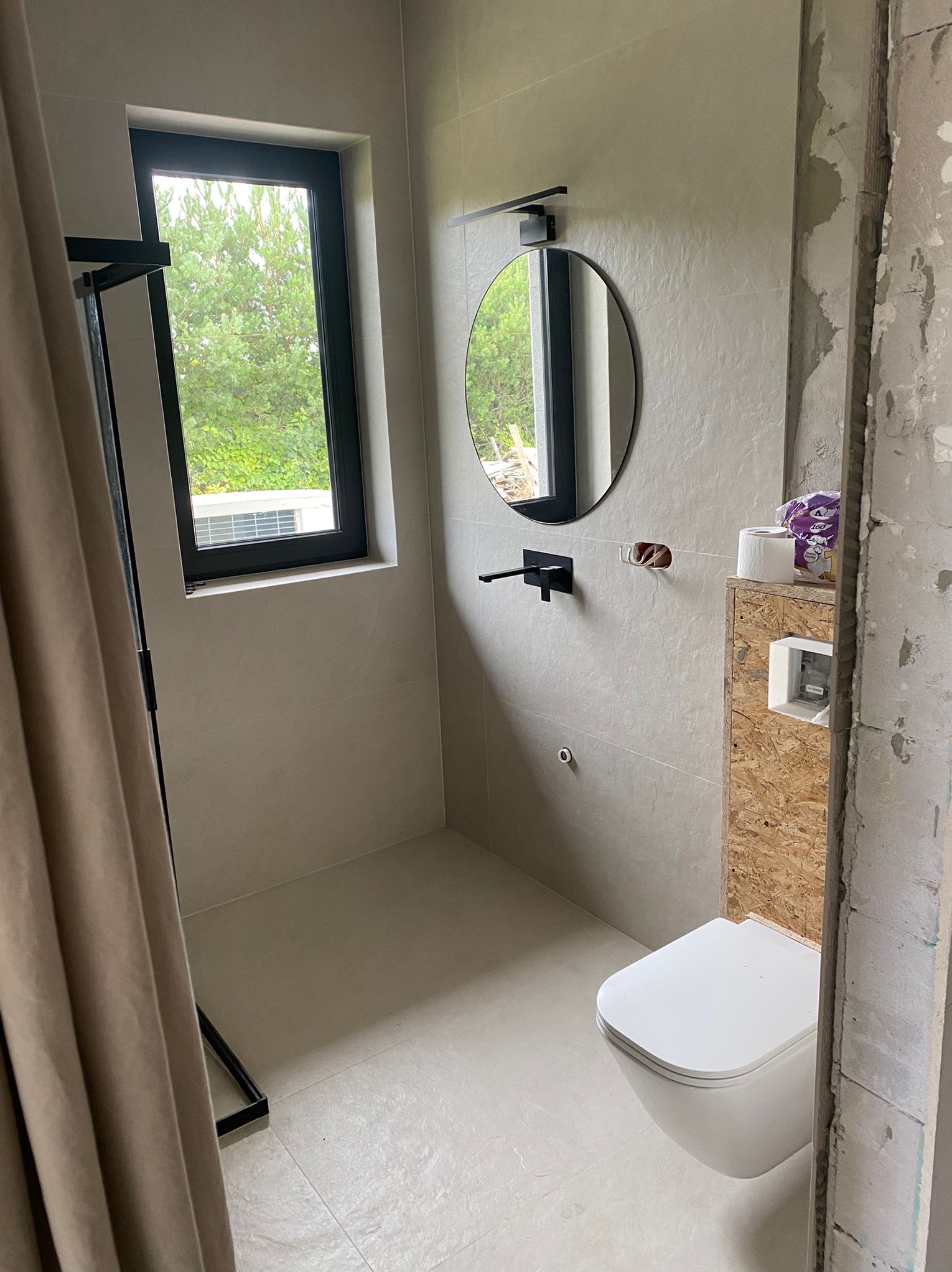 Jasna łazienka w trakcie wykańczania: szare płytki, okrągłe lustro z czarną ramą, wisząca toaleta. Widoczne niedokończone fragmenty ścian i instalacji.