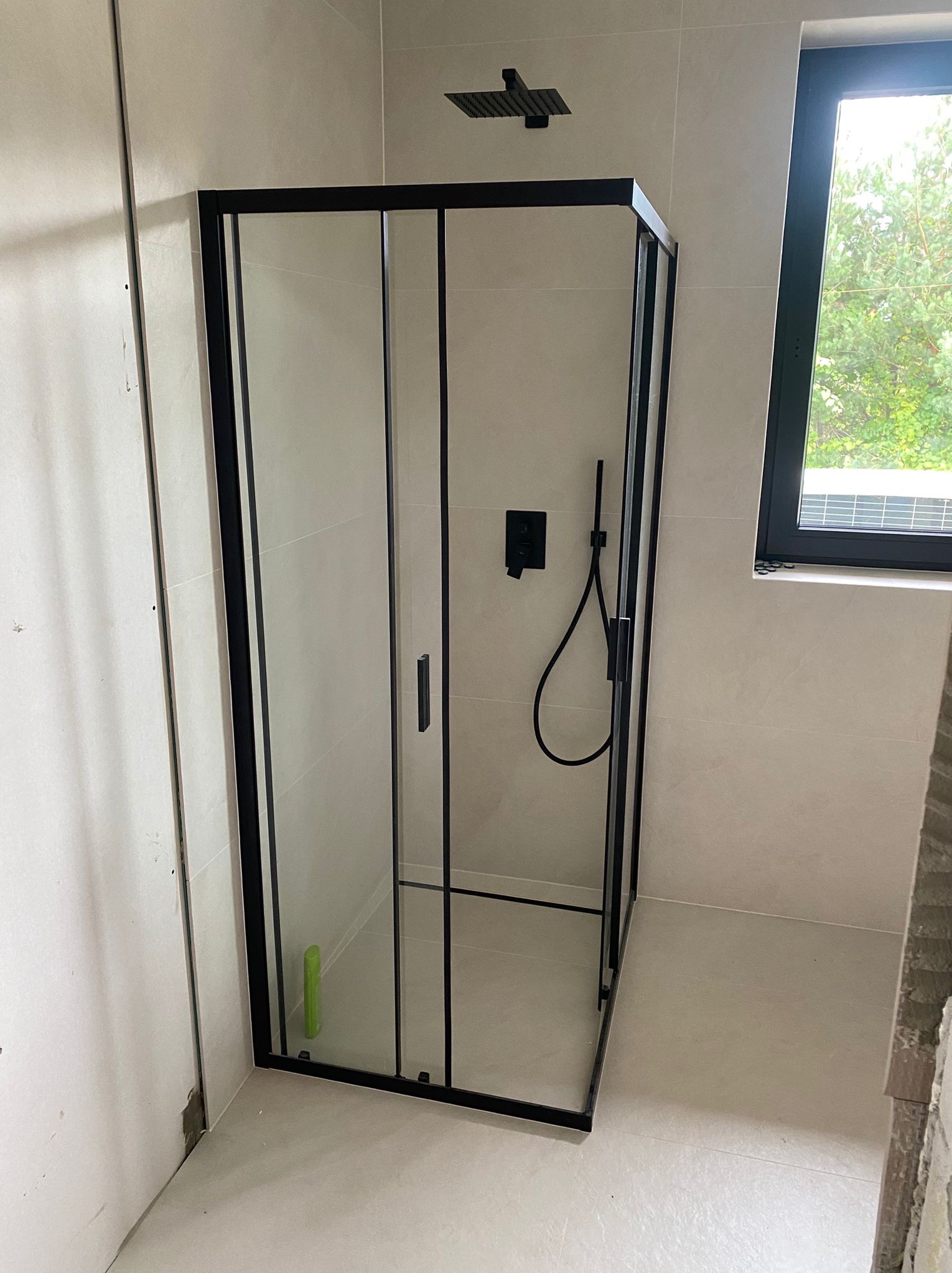 Nowoczesna kabina prysznicowa z czarnymi profilami i deszczownicą w wykończonej łazience z oknem. Widoczne płytki w jasnym odcieniu i fragment ściany w trakcie prac wykończeniowych.