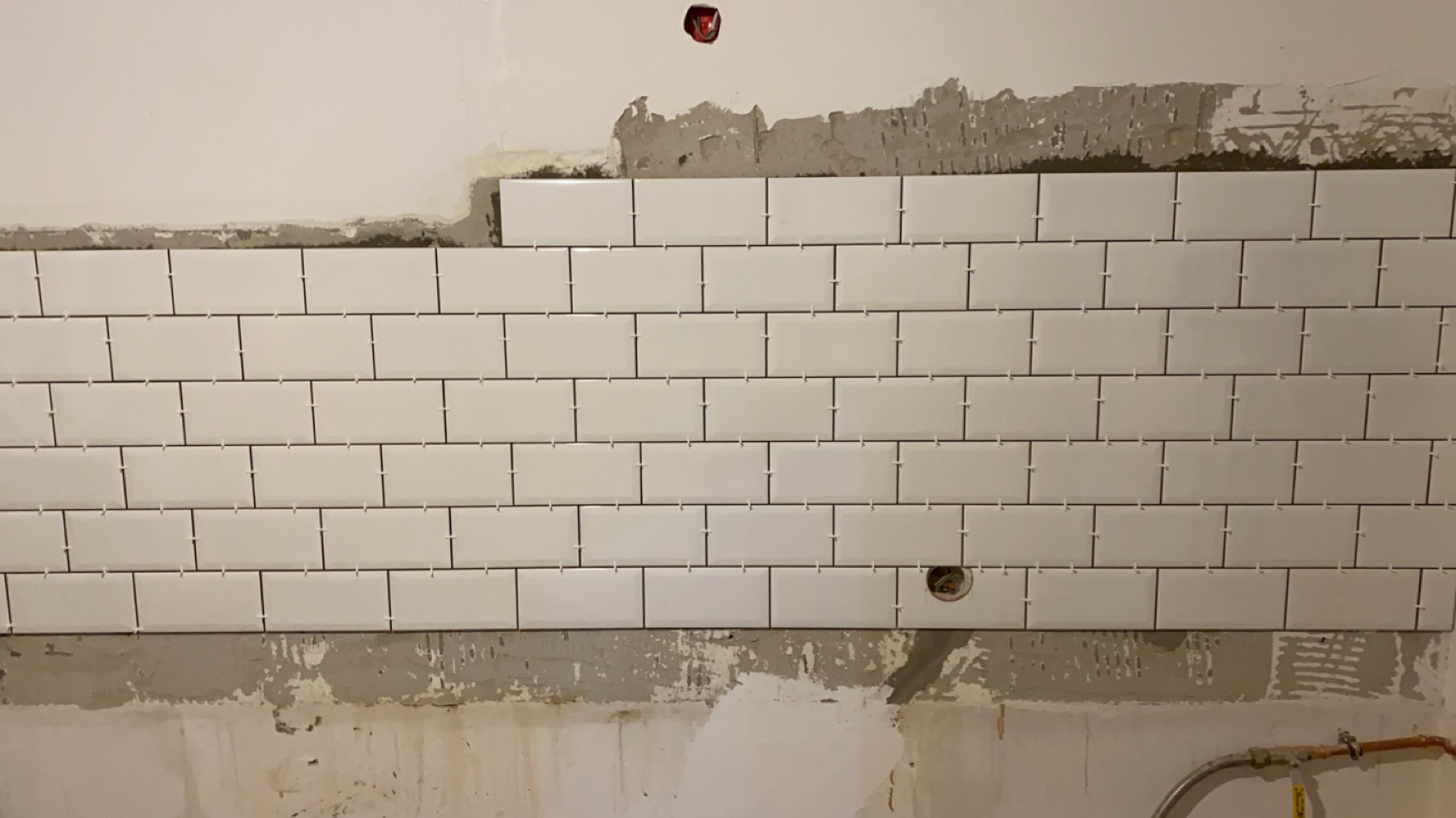 Układanie białych, prostokątnych płytek na ścianie z widocznymi krzyżykami dystansowymi i śladami zaprawy, w trakcie prac wykończeniowych.