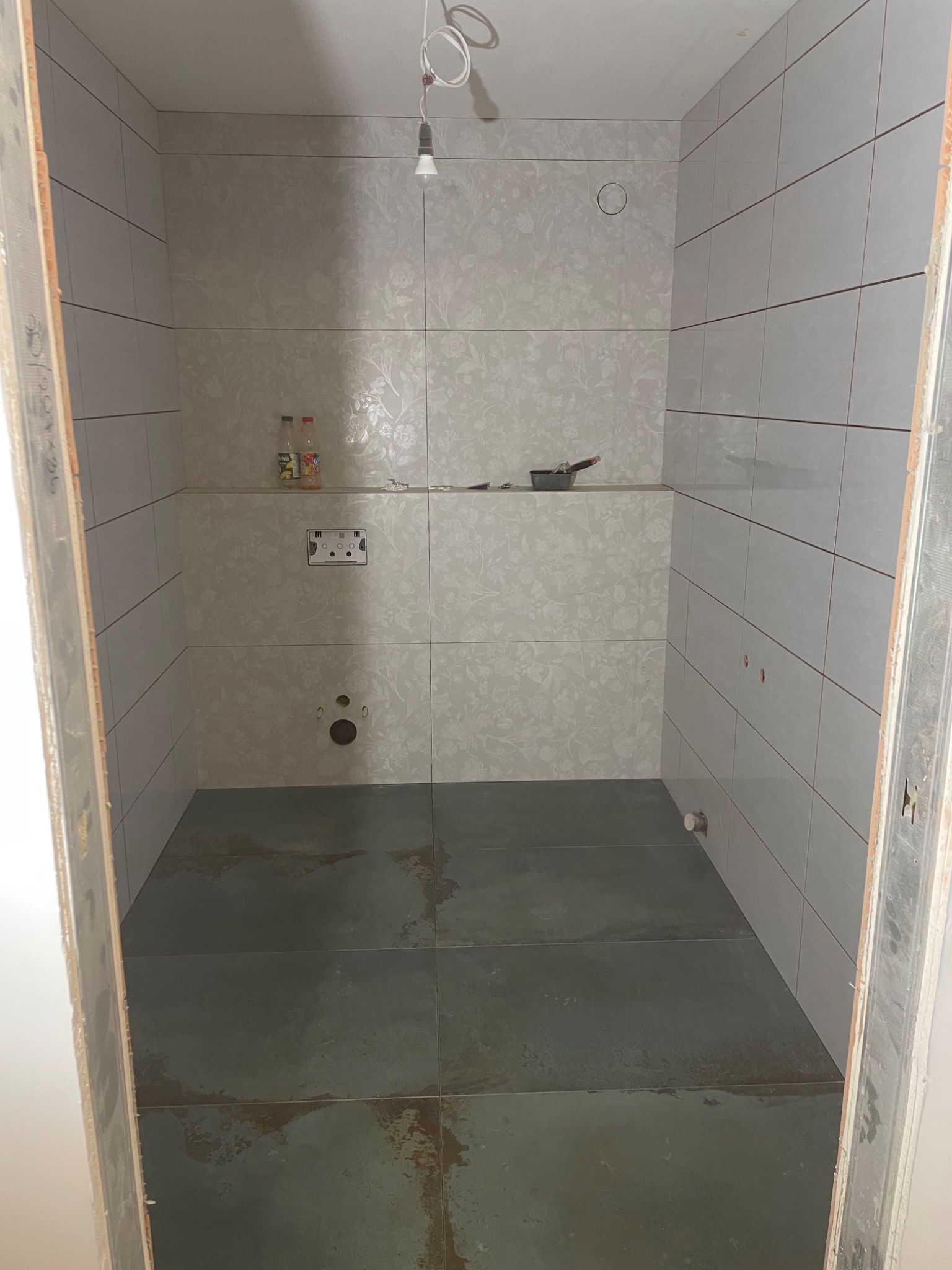 Wnętrze kabiny prysznicowej w trakcie wykańczania: ułożone płytki ścienne z delikatnym wzorem i ciemne płytki podłogowe. Widoczne narzędzia i materiały budowlane.