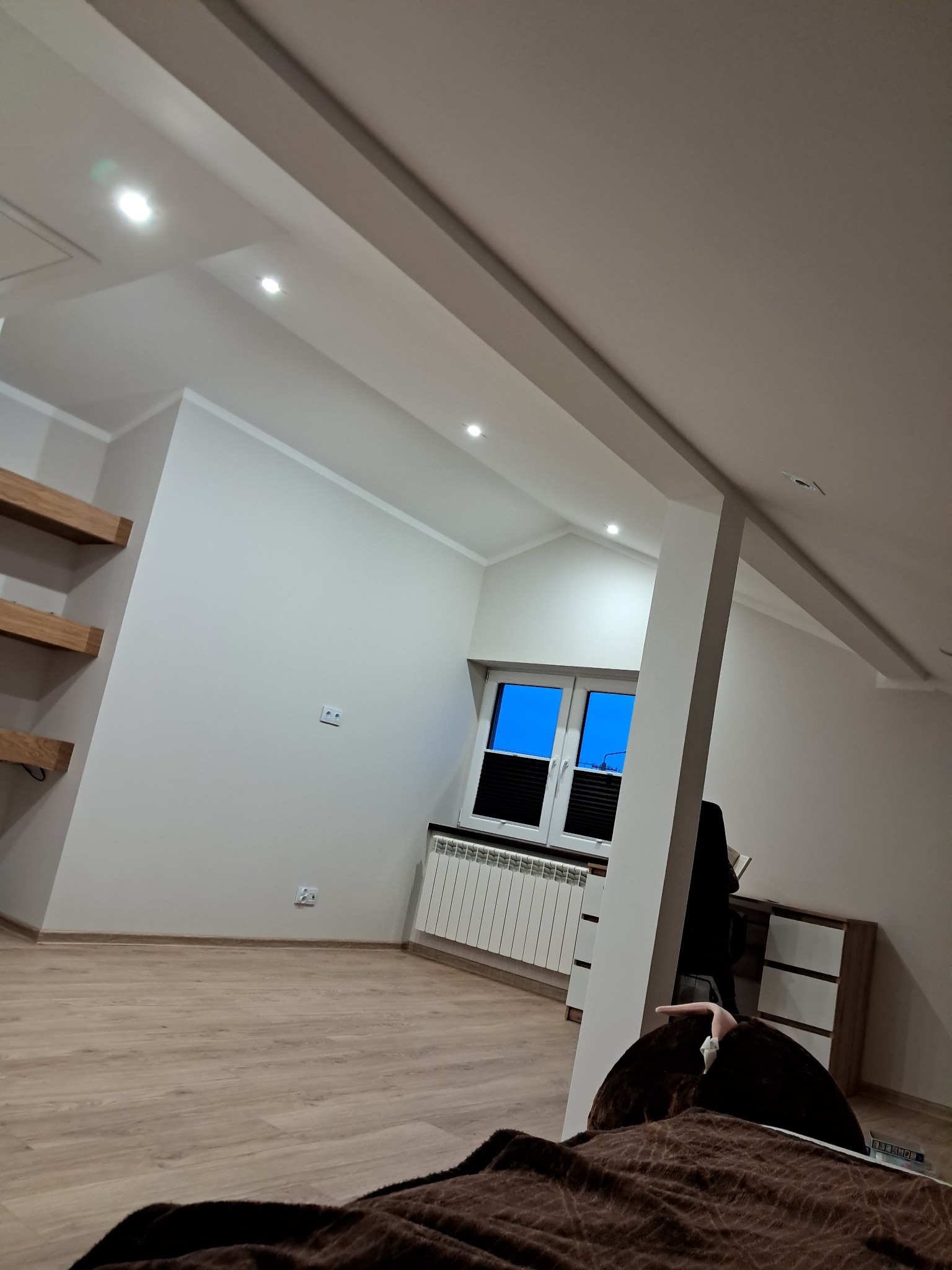 Jasne wnętrze poddasza z oknem, drewnianą podłogą i półkami. Widoczne skosy sufitu, białe ściany i grzejnik pod oknem. Minimalistyczny design.