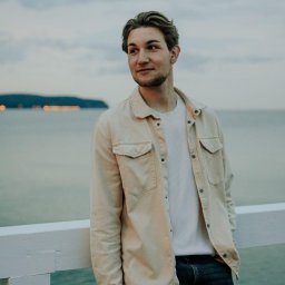 Tomasz Maćków - Młody mężczyzna w beżowej koszuli i białej koszulce, oparty o białą barierkę na tle morza i zachmurzonego nieba. Spogląda w lewo, w oddali widoczne światła miasta.