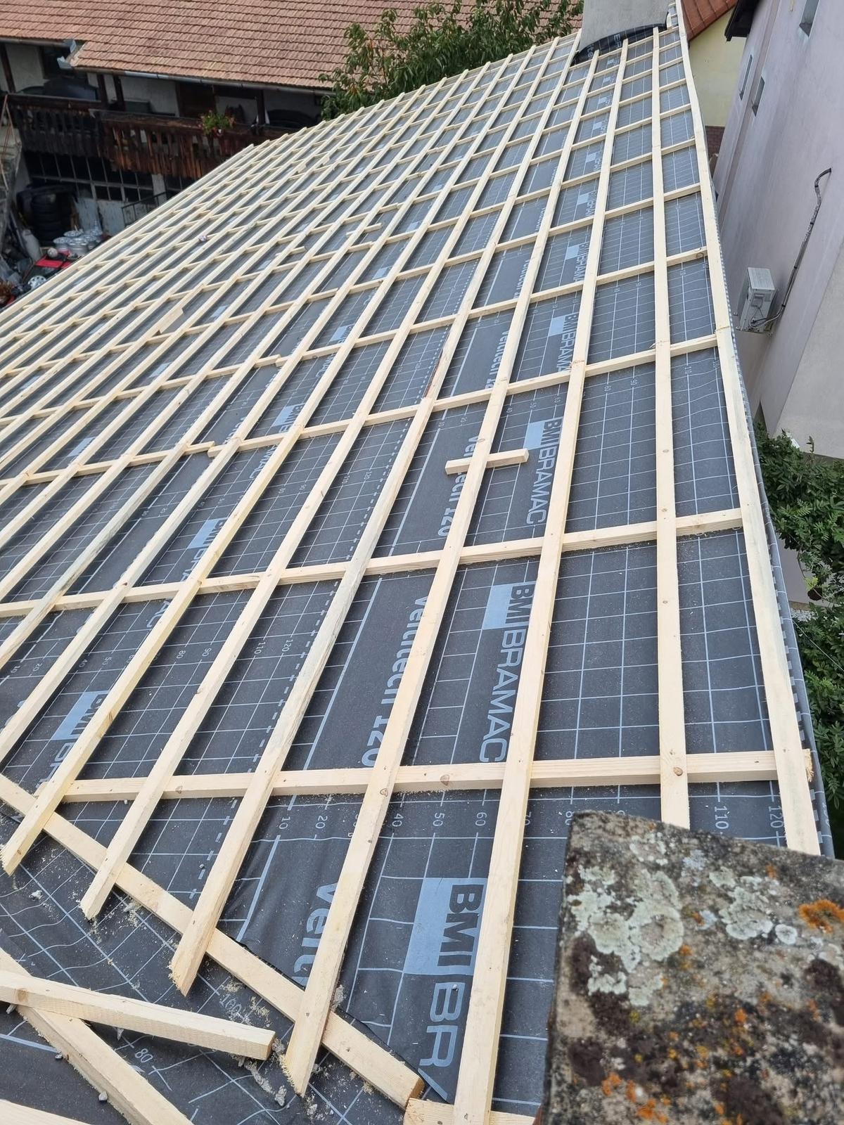 W trakcie montażu więźby dachowej na czarnej membranie z nadrukiem BMI Bramac. Widok z góry na konstrukcję z jasnego drewna, fragment starej dachówki w rogu.