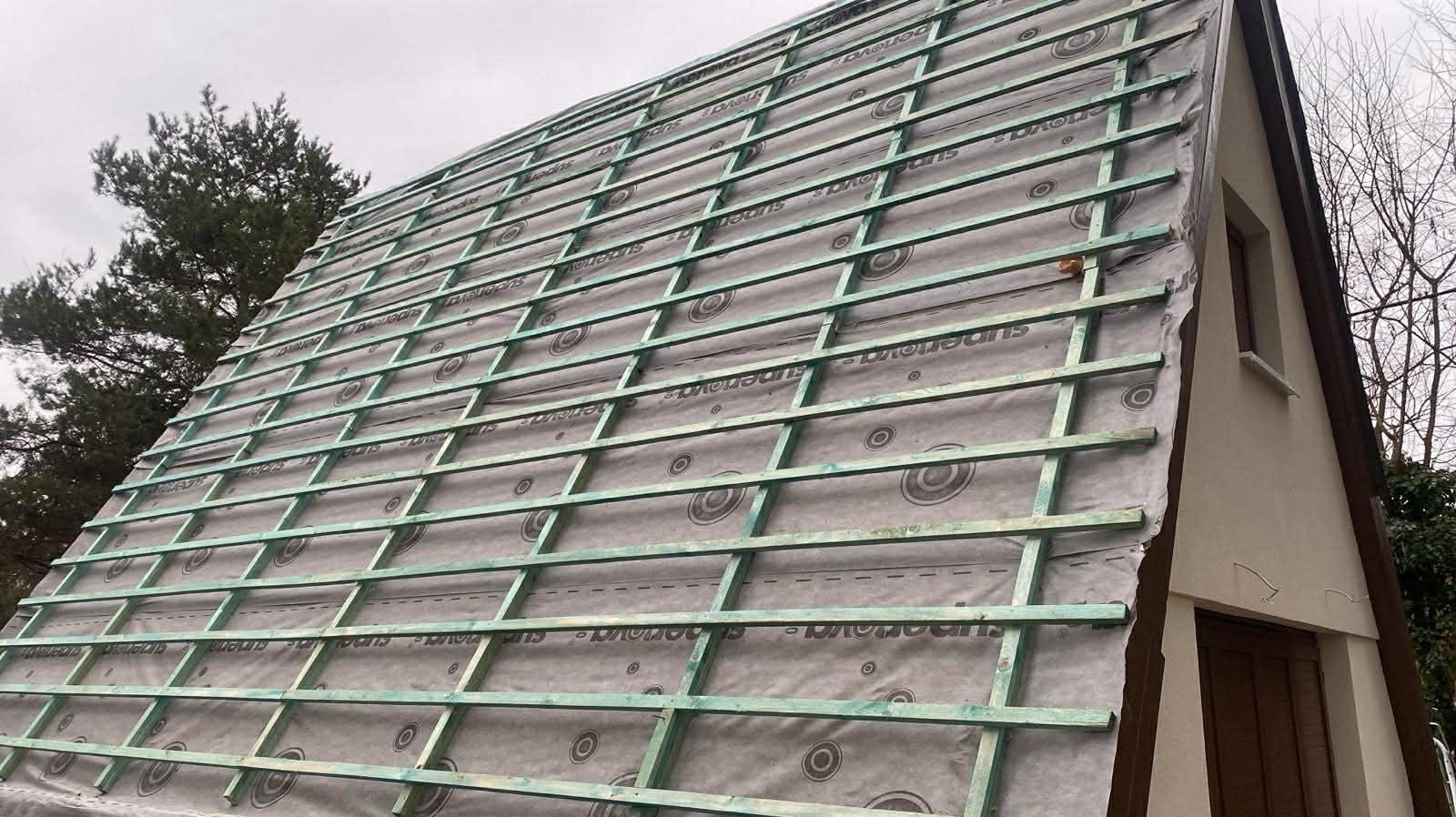 Konstrukcja dachu z łatami i membraną, widok z dołu pod kątem. Elementy drewniane w kolorze zielonym, tło stanowią drzewa i fragment budynku z oknem.