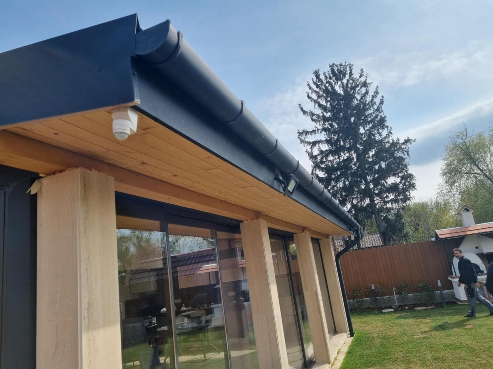 Nowoczesny dom z drewnianą podbitką dachową, czarnymi rynnami i kamerą. Widoczny fragment ogrodu i osoby w tle. Ujęcie z dołu, podkreślające detale wykończenia dachu.