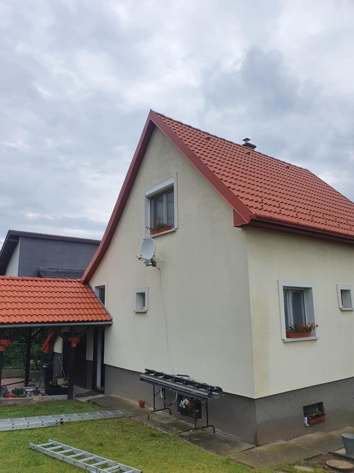 Dom jednorodzinny z nowym, czerwonym dachem z dachówki ceramicznej. Widoczne okna z kwiatami, drabina i stół z narzędziami dekarskimi na trawniku.