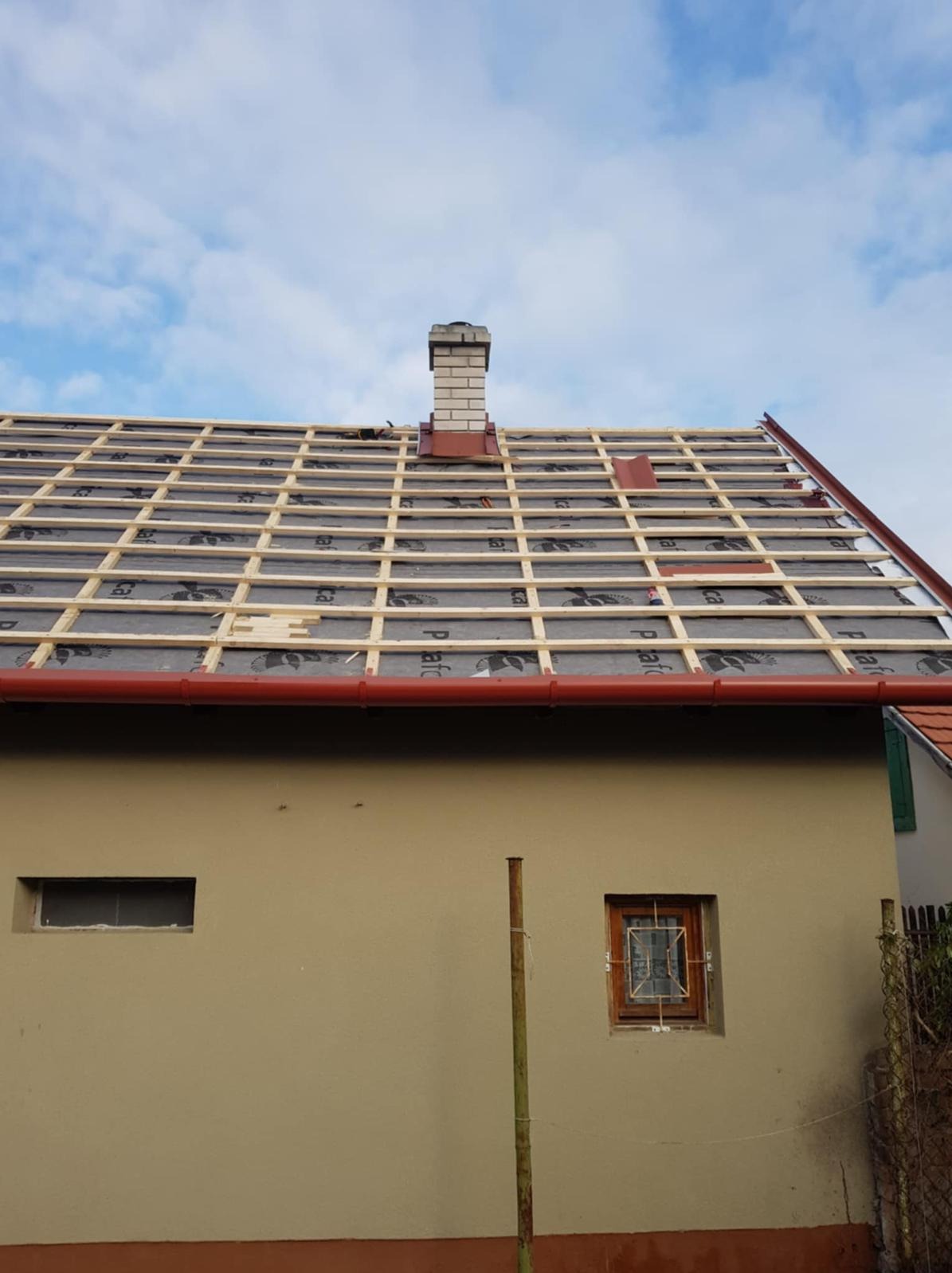 W trakcie prac: drewniana konstrukcja dachu z membraną, widoczny komin z cegły, czerwona rynna i fragment elewacji budynku. Częściowo ułożone dachówki.