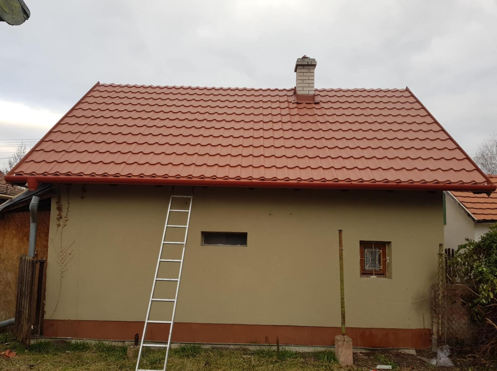 Dom z nowym, czerwonym dachem z blachodachówki i rynnami w tym samym kolorze. Aluminiowa drabina oparta o ścianę. Widok na komin i fragment ogrodu.