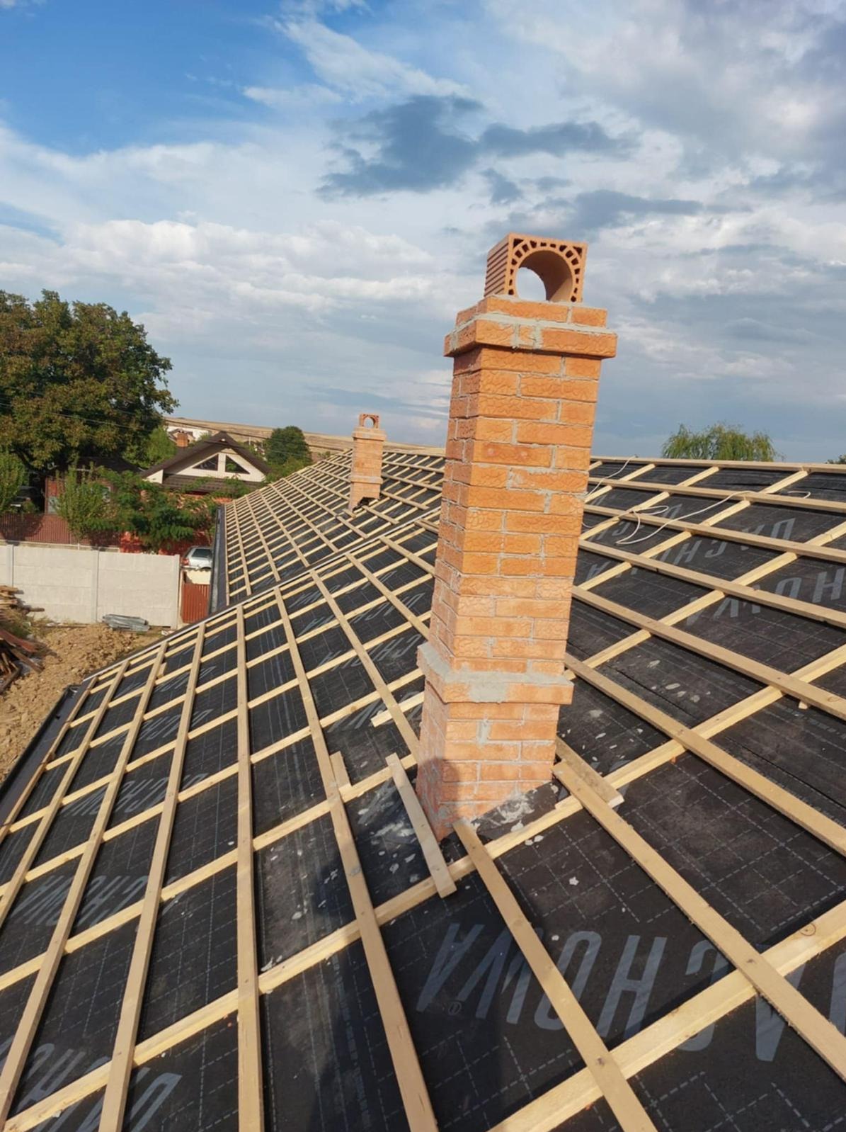 Dach z czarną membraną i drewnianą łatą, w tle komin z cegły. Widok z góry na konstrukcję dachu w trakcie prac dekarskich, z elementami murarskimi komina.