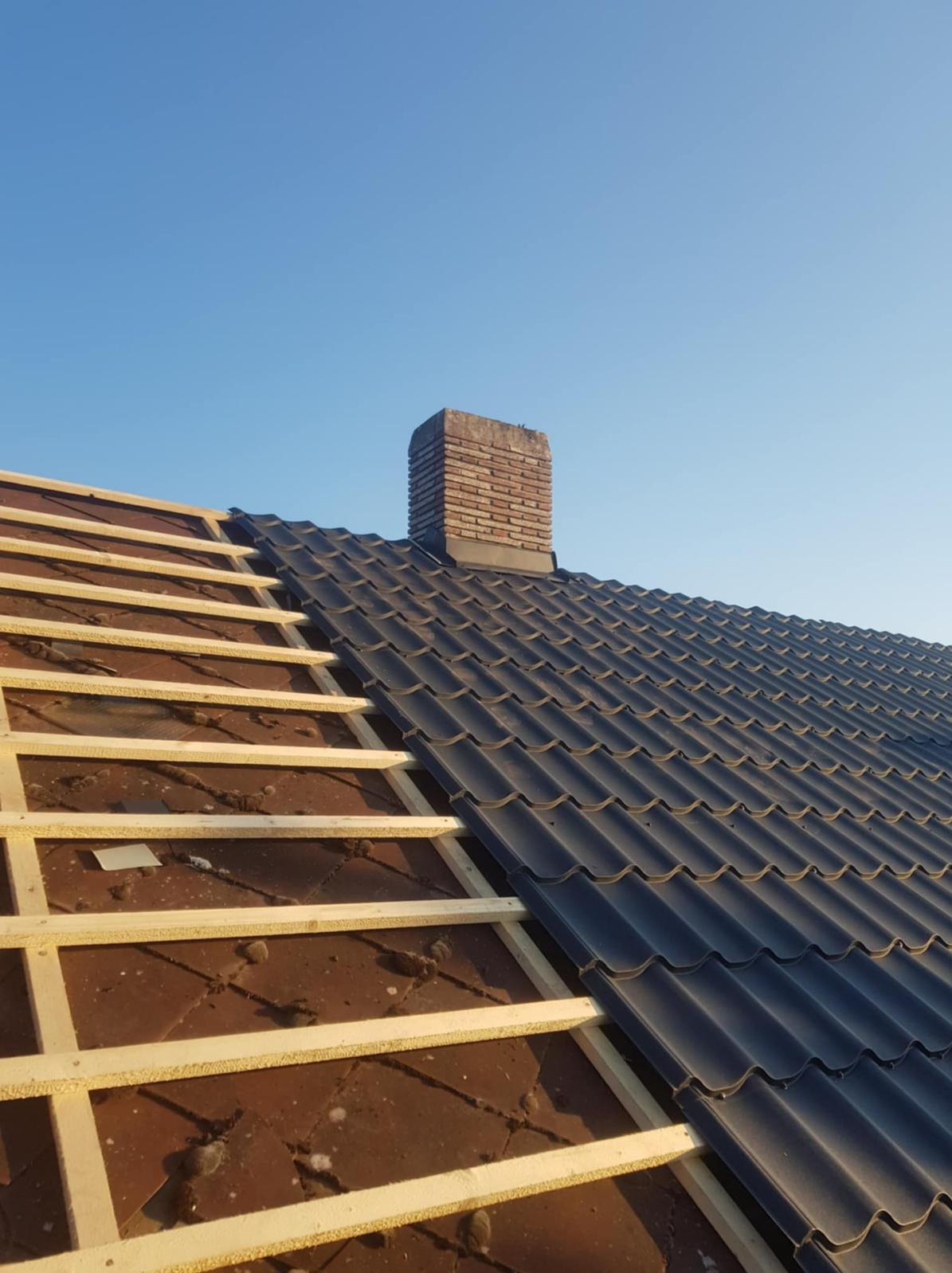 Częściowo pokryty dach z ciemnej blachy dachówkowej, widoczna więźba i komin na tle błękitnego nieba. Etap prac dekarskich.