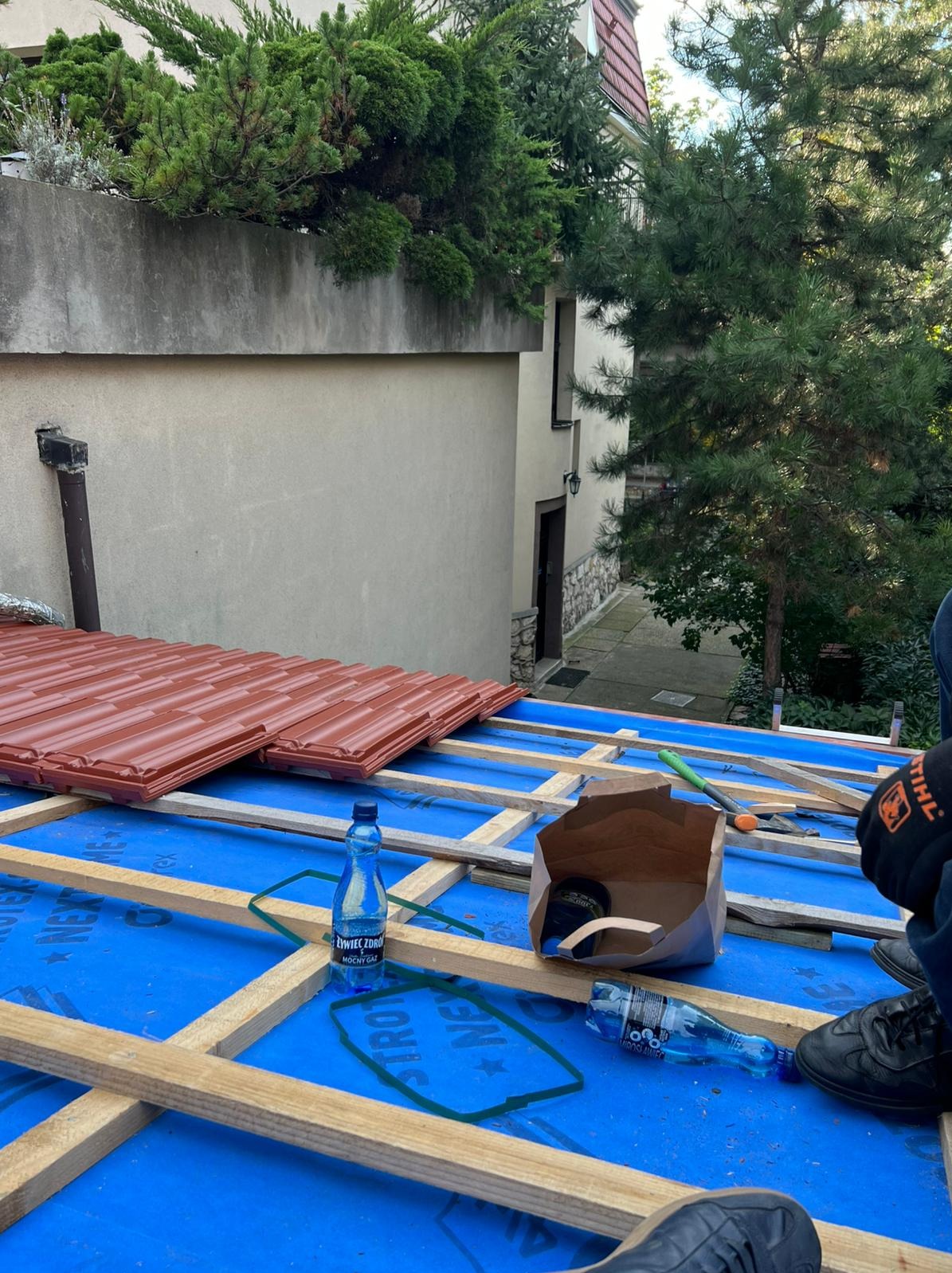 Montaż brązowej dachówki na niebieskiej membranie dachowej, widoczne drewniane łaty, narzędzia i butelki wody. Praca w toku, częściowo widoczny budynek w tle.
