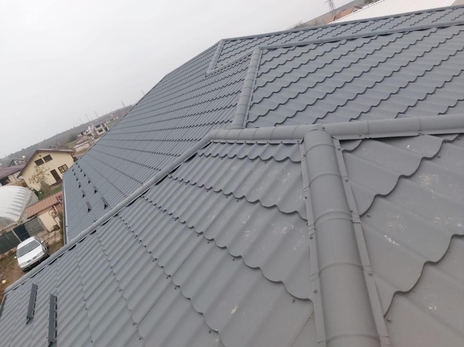 Szary dach z blachy trapezowej z rynnami w kolorze, widok z góry na tle zabudowy jednorodzinnej w pochmurny dzień. Charakterystyczne mocowania rynien.