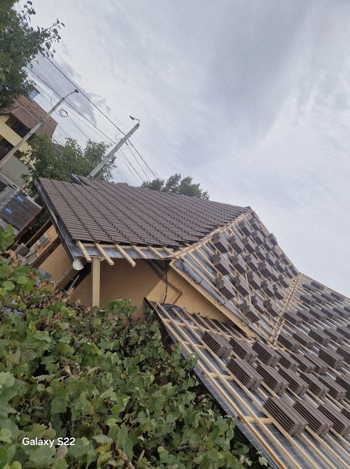 Układanie brązowej dachówki na drewnianej konstrukcji dachu domu jednorodzinnego, widok z dołu, częściowo zasłonięty przez zieloną roślinność.