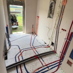 Hydrokom - Instalacja wodna i elektryczna w nowym budynku: rury czerwone i niebieskie na podłodze, kable w ścianie, pianka montażowa. Widok na zewnątrz przez drzwi.