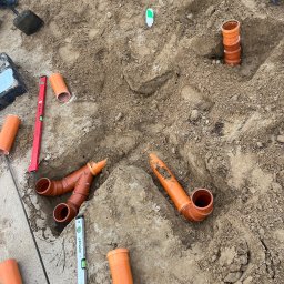 Hydrokom - Pomarańczowe rury kanalizacyjne ułożone w wykopie ziemnym, widoczne poziomice i inne akcesoria instalacyjne. Etap prac ziemnych przed zasypaniem.