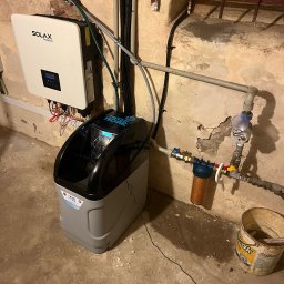 Hydrokom - Szara stacja uzdatniania wody Water Technic z podłączonym filtrem i licznikiem, obok biały inwerter Solax Power na tle ściany z rurami i oknem z kratą.