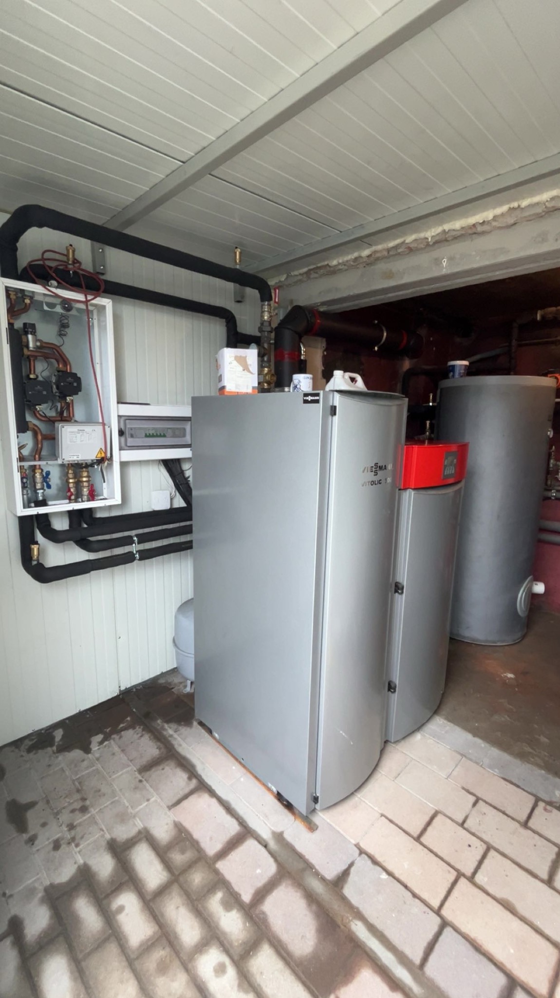 Nowoczesna kotłownia z szarym piecem Viessmann Vitolic 100, instalacją hydrauliczną w białej szafce i zasobnikiem wody. Podłoga z szarej kostki brukowej.