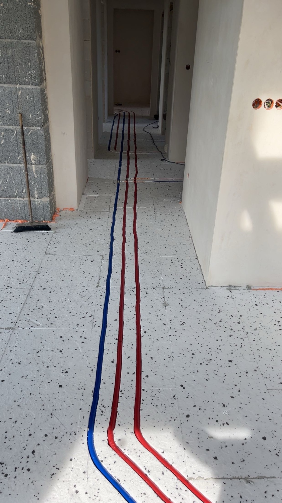 Ułożone rury instalacji wodnej (czerwone i niebieskie) na podłodze w surowym wnętrzu, widok z perspektywy, z widocznymi puszkami elektrycznymi w ścianie.