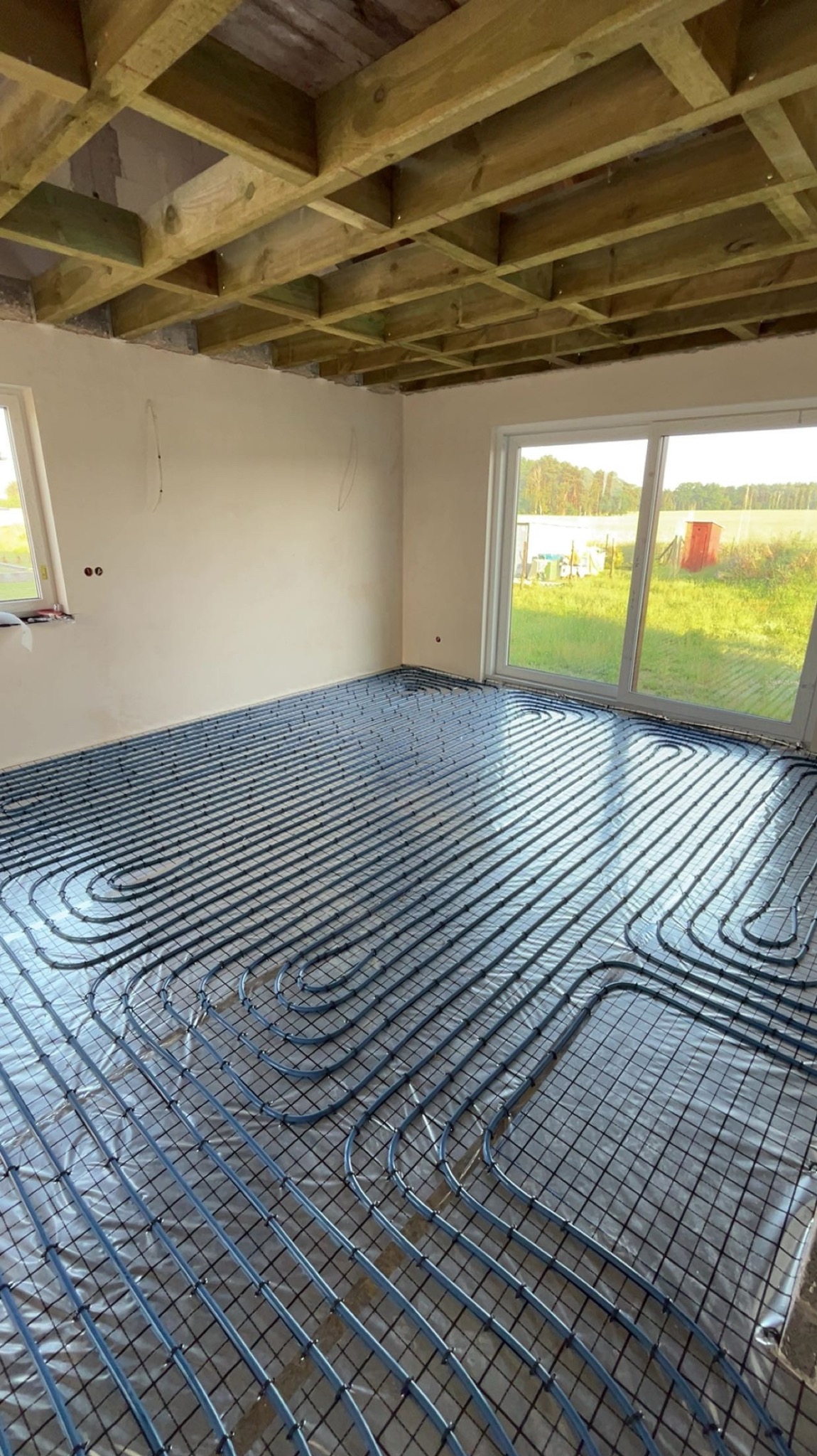 Ułożone rury ogrzewania podłogowego na folii, widoczne okna i surowy sufit z drewnianych belek. Etap prac przed wylaniem posadzki.