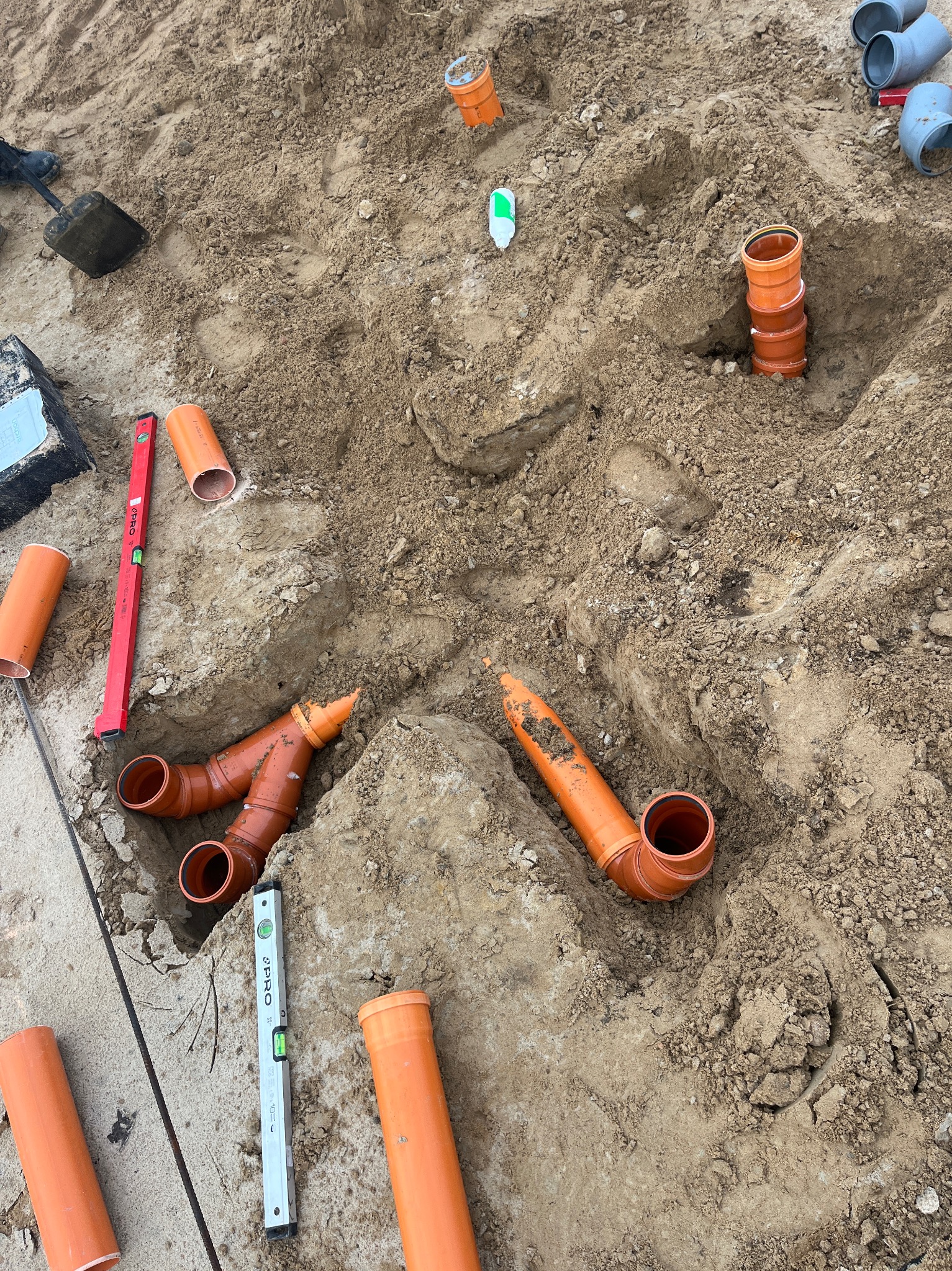 Pomarańczowe rury kanalizacyjne ułożone w wykopie ziemnym, widoczne poziomice i inne akcesoria instalacyjne. Etap prac ziemnych przed zasypaniem.