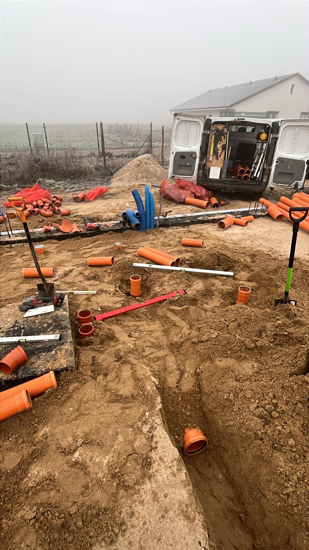 Wykop z pomarańczowymi rurami kanalizacyjnymi, poziomice, łopaty i samochód dostawczy w tle. Prace ziemne w trakcie realizacji w pochmurny dzień.
