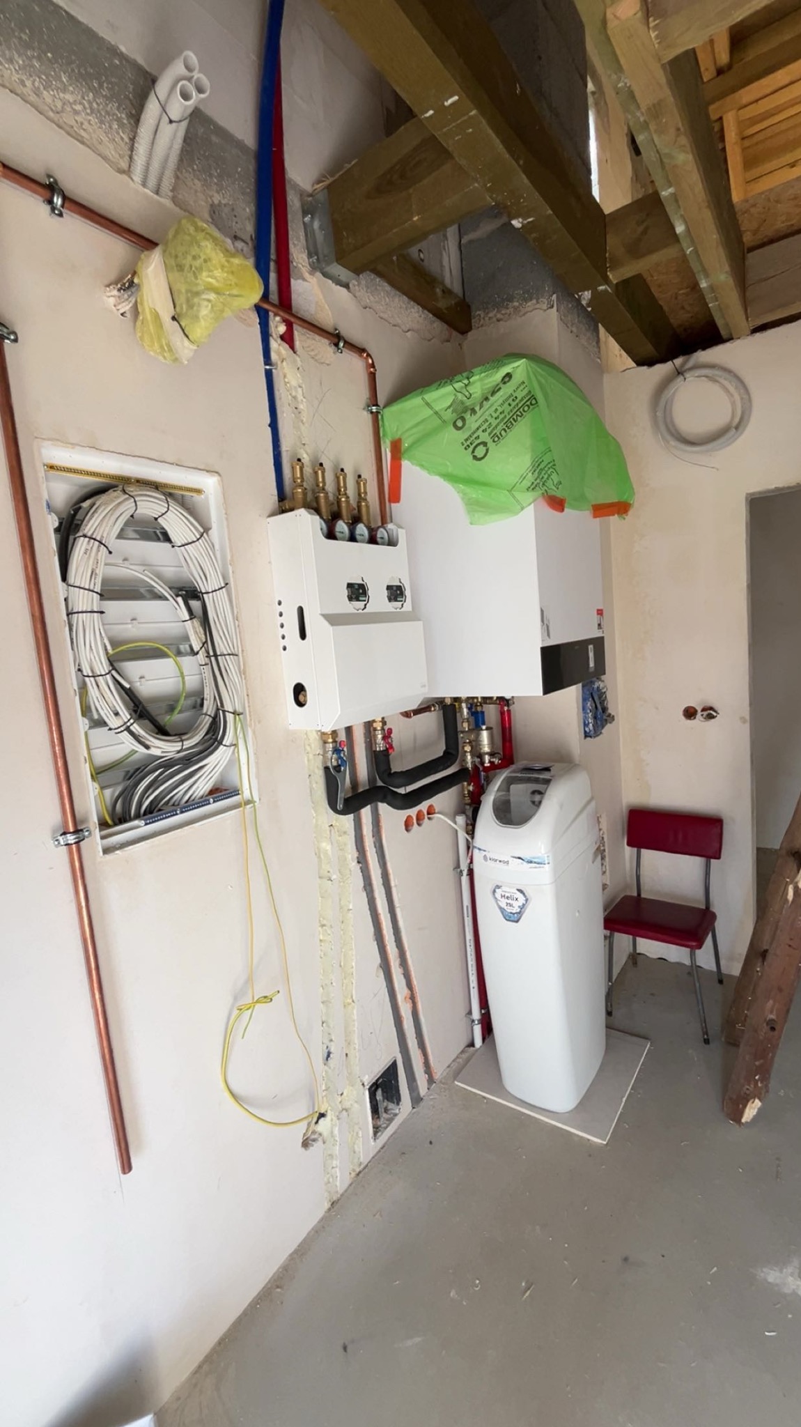 Biały piec grzewczy z instalacją hydrauliczną i zmiękczaczem wody w surowym wnętrzu. Widoczne miedziane rury, kable w ścianie i drewniana konstrukcja stropu.