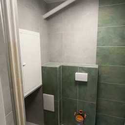 Dawid Kos - Wykończona łazienka z zielonymi i szarymi płytkami, podwieszana toaleta i biała szafka rewizyjna. Widoczny fragment rury pod sufitem i okrągła lampa.