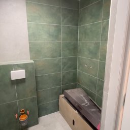 Dawid Kos - Wnętrze łazienki w trakcie remontu: zielone płytki na ścianach, przygotowane miejsce na toaletę, karton z armaturą. Widoczne elementy instalacji.