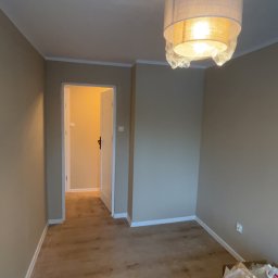 Dawid Kos - Pokój po malowaniu ścian na beżowo, z nową podłogą imitującą drewno i lampą pod sufitem. Widoczne otwarte drzwi do innego pomieszczenia i materiały budowlane w rogu.