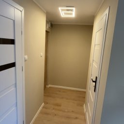 Dawid Kos - Wykończony korytarz z drewnianą podłogą, białymi drzwiami z czarnymi akcentami i nowoczesnym oświetleniem sufitowym. Ściany w odcieniach beżu i szarości.