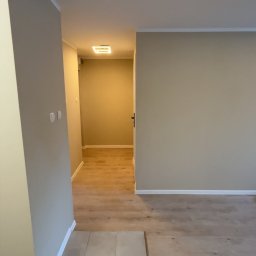 Dawid Kos - Beżowe ściany po malowaniu w pustym pokoju z fragmentem ułożonej podłogi z płytek i drewnianych paneli. Widok na korytarz z oświetleniem sufitowym.