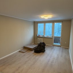 Dawid Kos - Pokój po remoncie podłogi z panelami, widoczne worki z odpadami i kartony po panelach. Ściany pomalowane na beżowo, okno balkonowe z widokiem na zewnątrz.