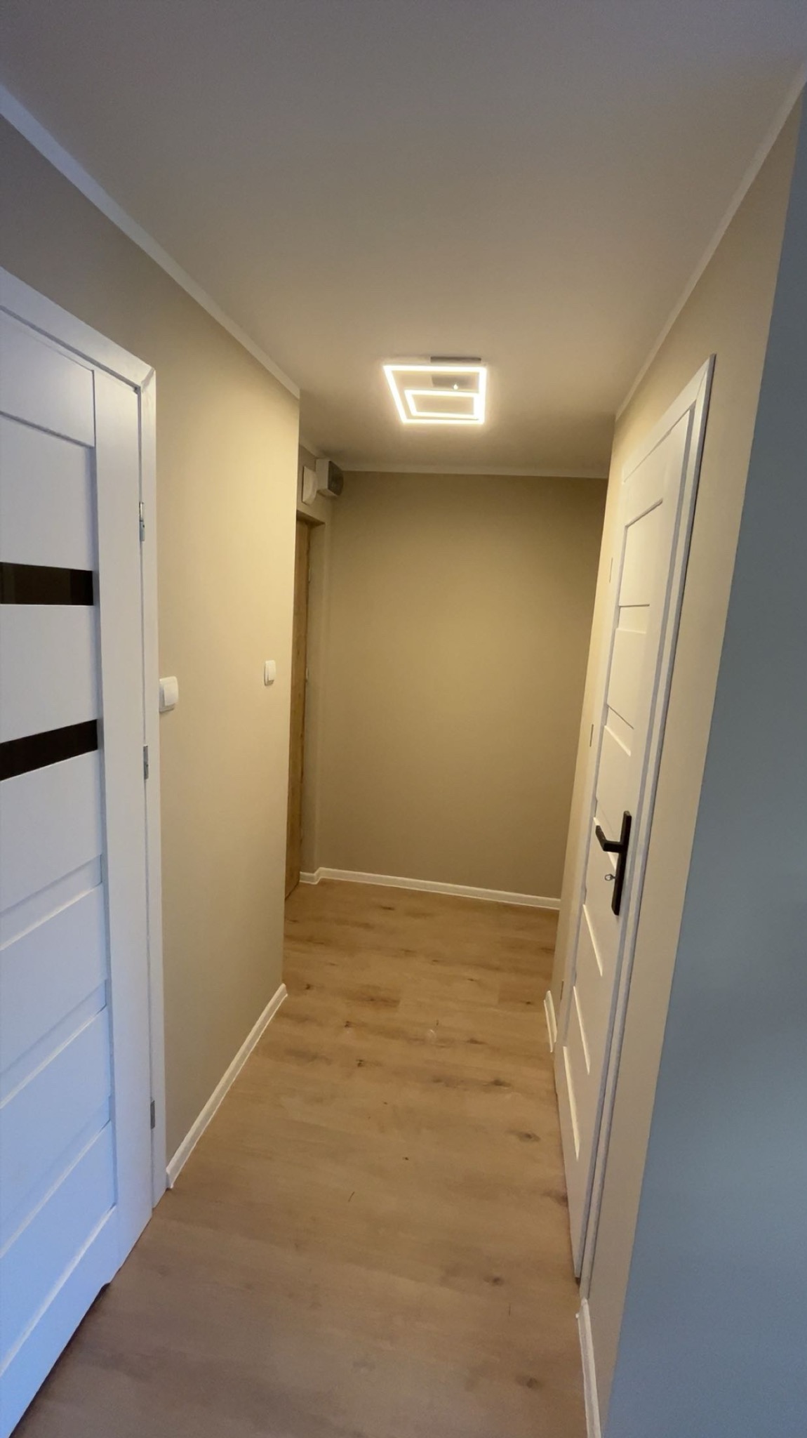 Wykończony korytarz z drewnianą podłogą, białymi drzwiami z czarnymi akcentami i nowoczesnym oświetleniem sufitowym. Ściany w odcieniach beżu i szarości.