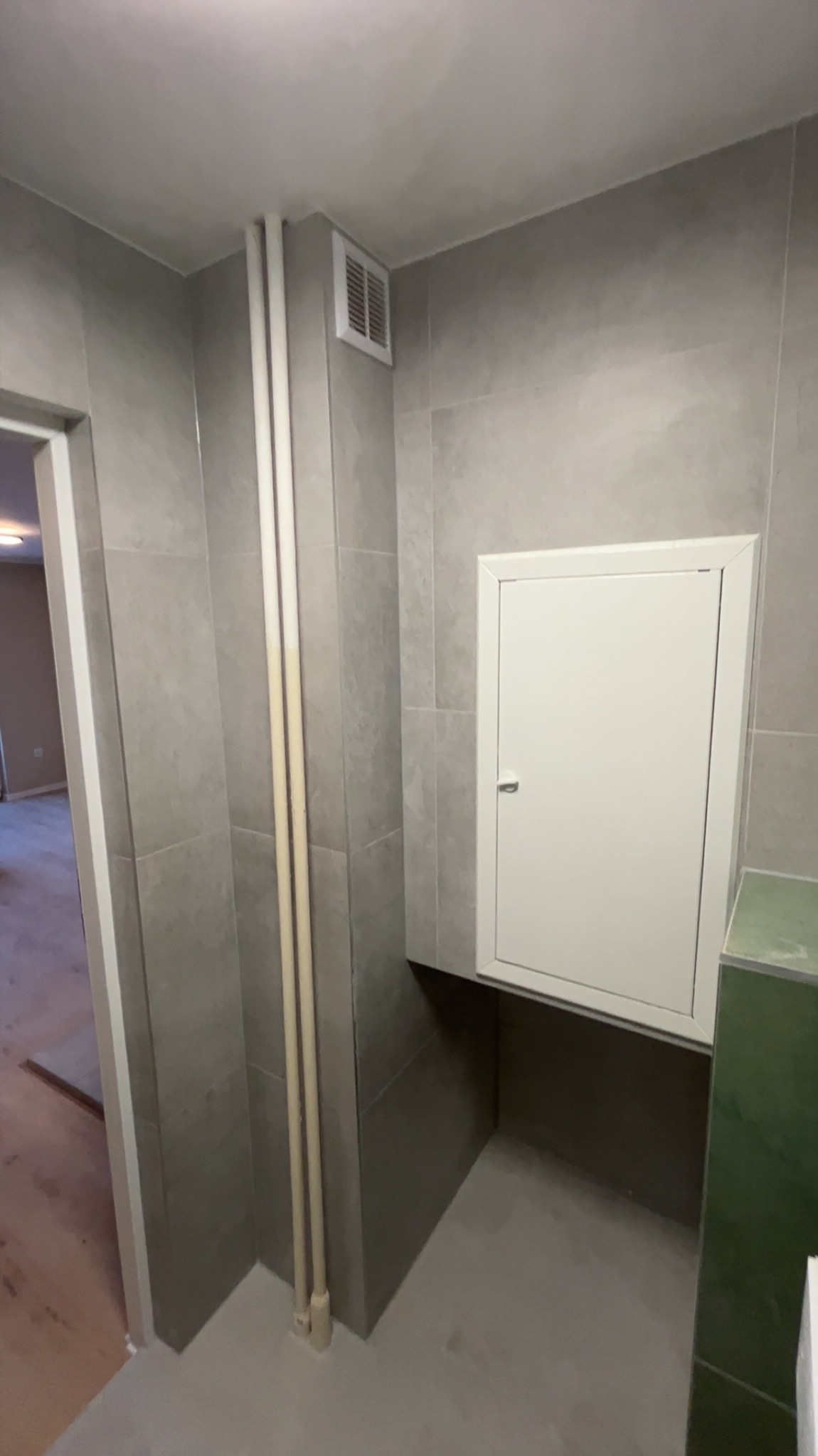 Szara łazienka po remoncie z ukrytą szafką rewizyjną, widoczne rury i kratka wentylacyjna. Minimalistyczny design, płytki imitujące kamień.