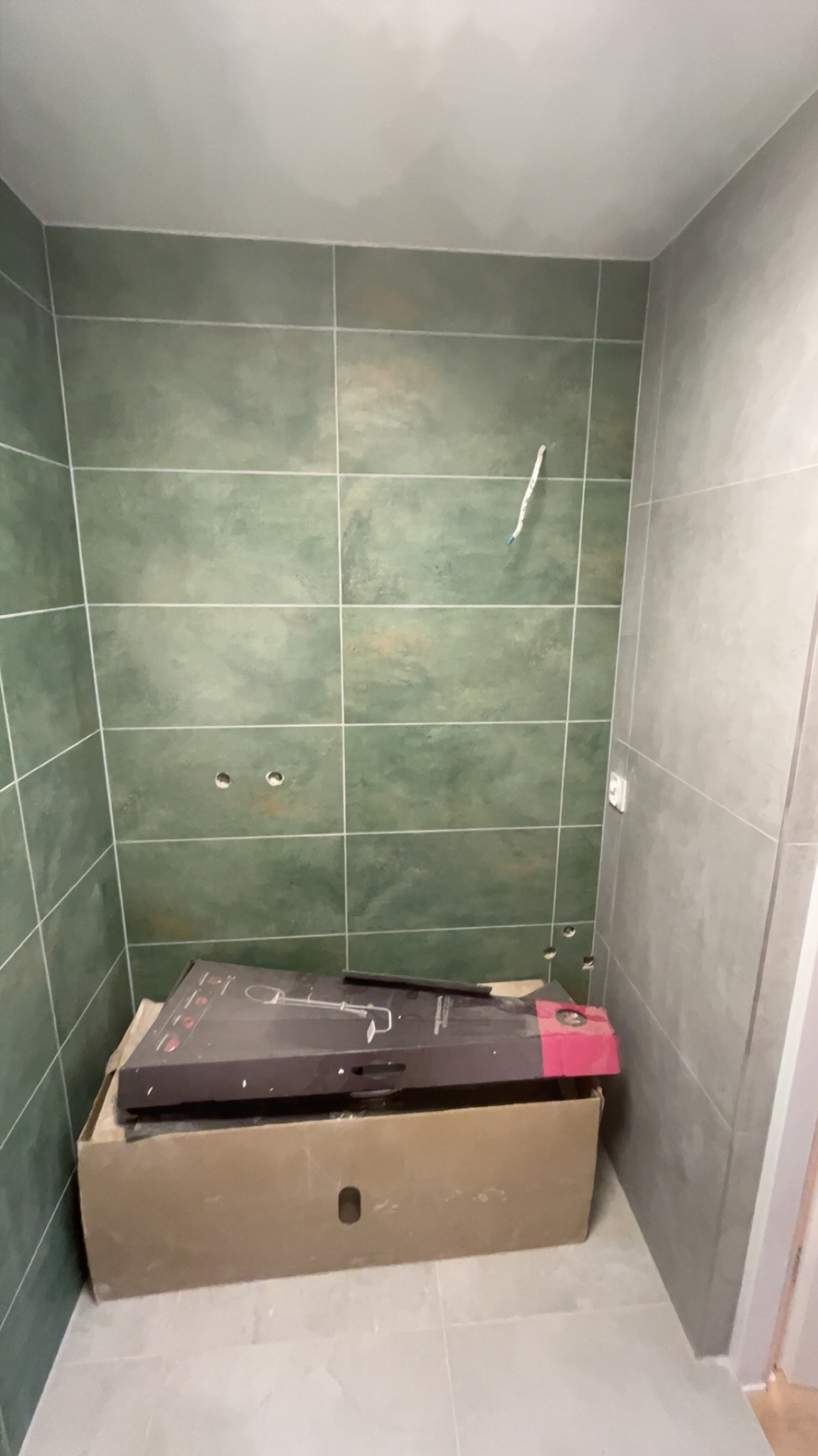 Przygotowanie do montażu armatury w łazience w Bogorii. Widoczne zielone płytki i karton z nową baterią prysznicową.