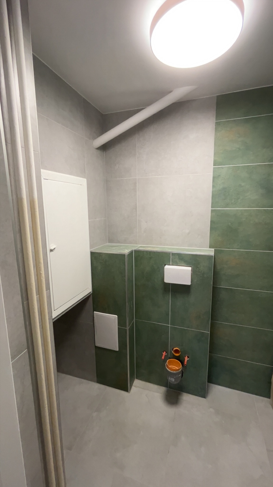 Wykończona łazienka z zielonymi i szarymi płytkami, podwieszana toaleta i biała szafka rewizyjna. Widoczny fragment rury pod sufitem i okrągła lampa.
