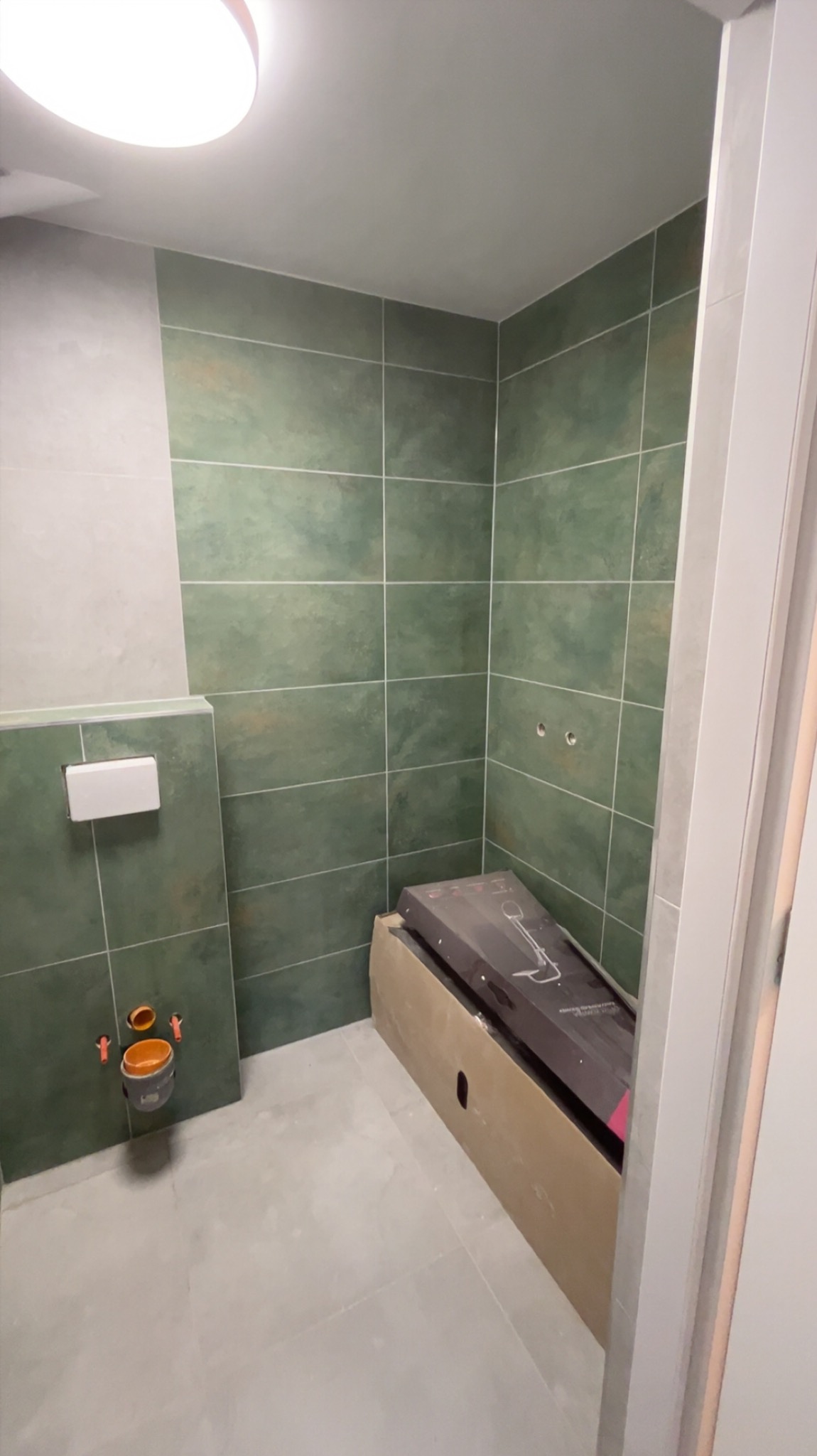 Wnętrze łazienki w trakcie remontu: zielone płytki na ścianach, przygotowane miejsce na toaletę, karton z armaturą. Widoczne elementy instalacji.