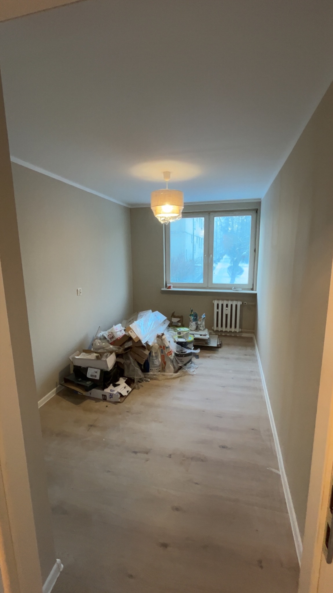 Pokój z panelami podłogowymi w odcieniach szarości, beżowe ściany, okno z grzejnikiem i sterta kartonów po remoncie. Lampa sufitowa włączona.