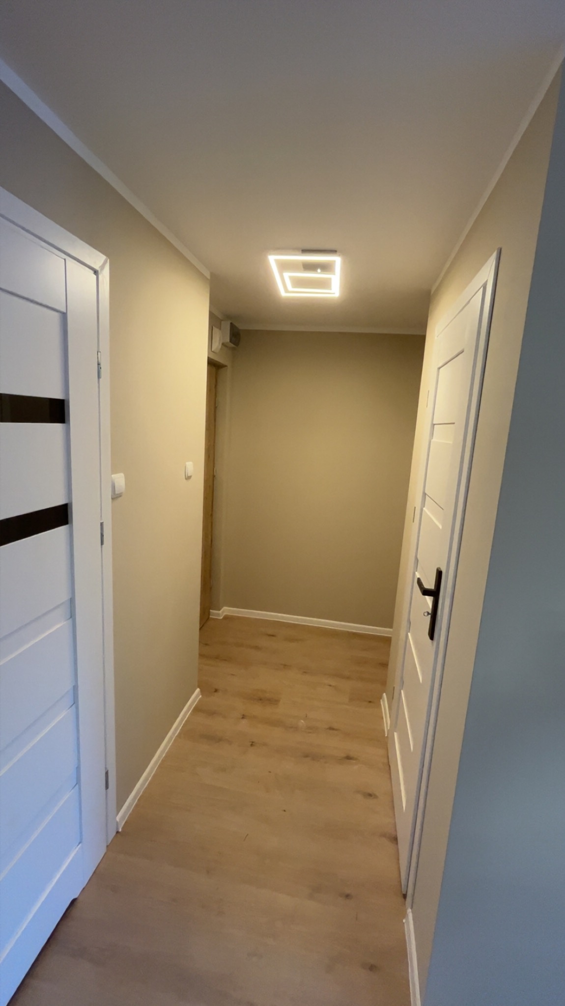 Wykończony korytarz z drewnianą podłogą, białymi drzwiami z czarnymi akcentami i nowoczesnym oświetleniem sufitowym. Ściany w odcieniach beżu i szarości.